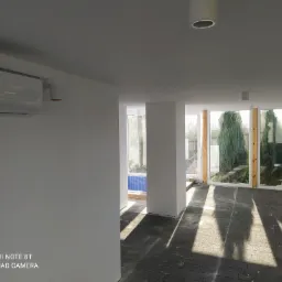 Jasne, przestronne wnętrze z dużymi oknami i szarą podłogą z kostki, widoczny klimatyzator na ścianie i wnęka z niebieską mozaiką w oddali.