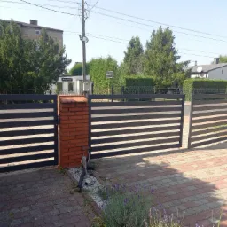 Nowoczesna brama wjazdowa z poziomymi, czarnymi przęsłami, osadzona na ceglanych słupkach, z widoczną kostką brukową i zielenią w tle.