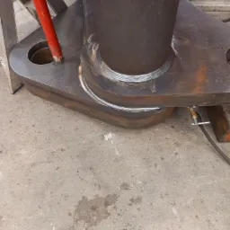 Metalowy element konstrukcyjny po spawaniu, widoczne spoiny, rura wtopiona w grubą płytę z otworami, szary beton na podłodze.