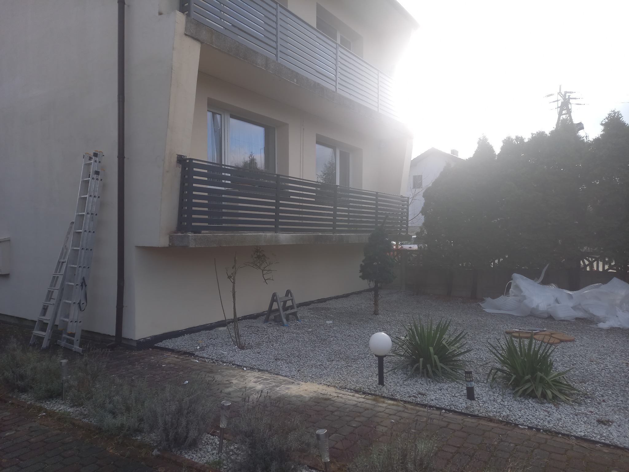 Nowoczesny dom z szarym poziomym ogrodzeniem balkonowym i panelowym ogrodzeniem wokół posesji, widoczne z zewnątrz, z drabina opartą o ścianę.