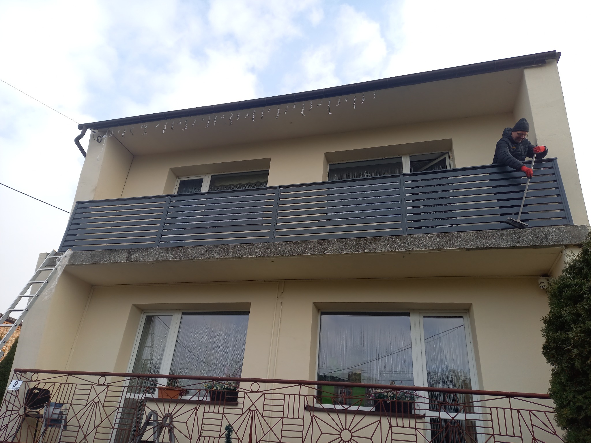Mężczyzna w kurtce i czapce zamiata liście z balkonu z szarą, poziomą balustradą. W tle budynek z beżową elewacją i drabina oparta o ścianę.