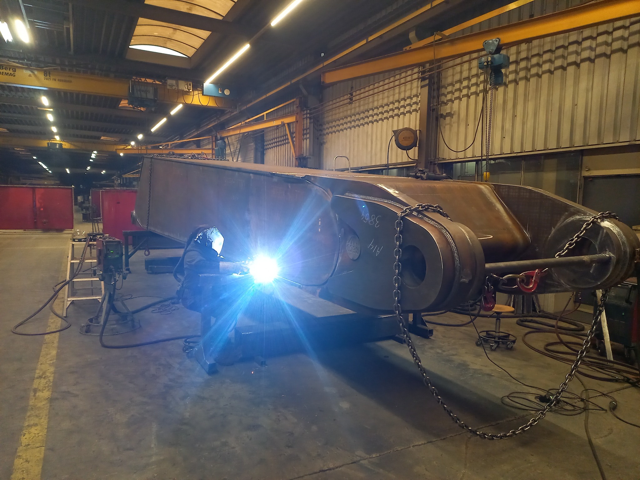 Spawanie elementu konstrukcyjnego w hali przemysłowej; widoczny spawacz w masce ochronnej, iskry, łańcuchy, suwnica pod stropem.