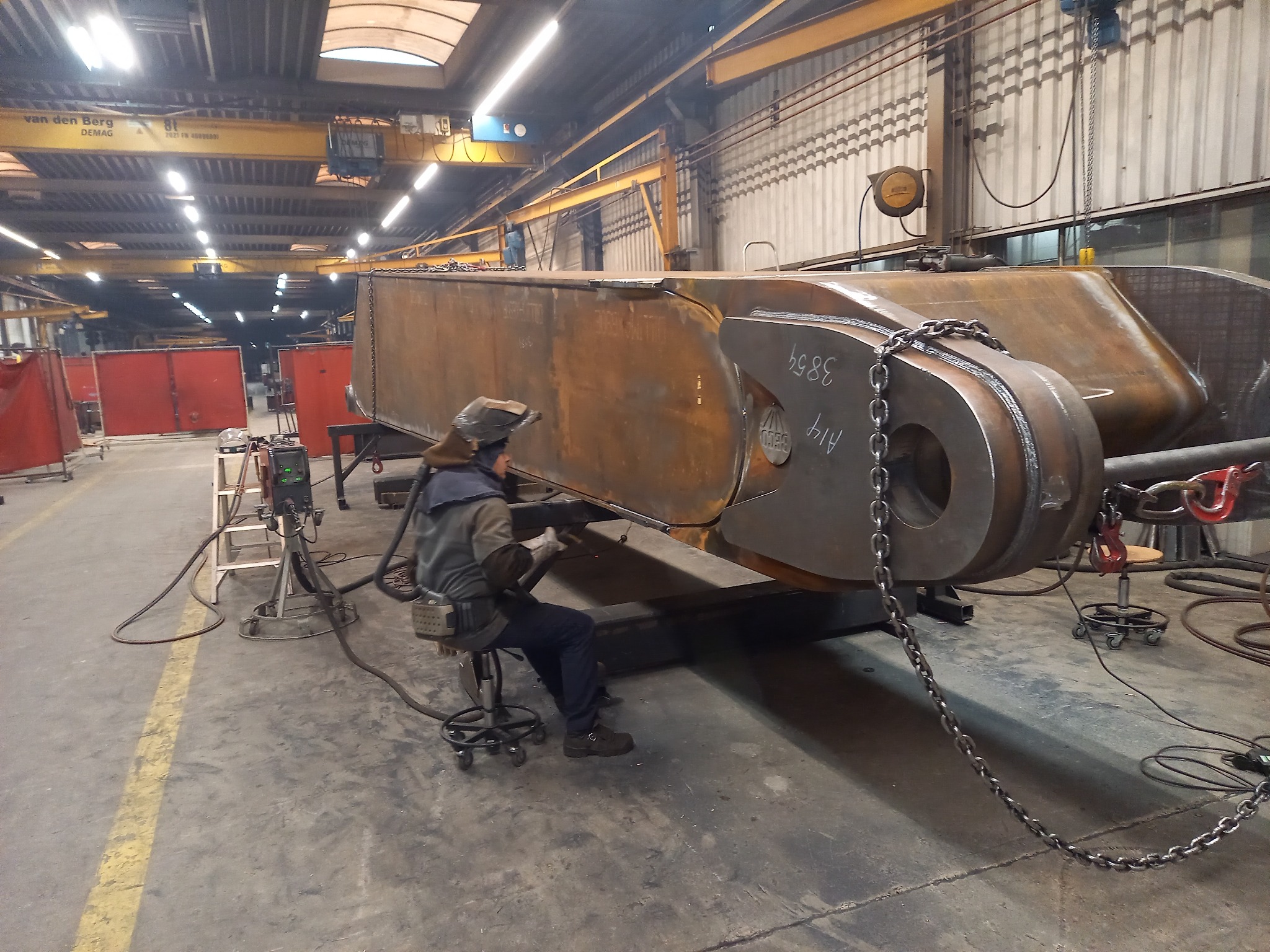 Spawacz w kombinezonie i masce spawalniczej, siedzący na krześle na kółkach, spawa duży, metalowy element konstrukcyjny w hali produkcyjnej, widoczny sprzęt spawalniczy i łańcuchy zabezpieczające.