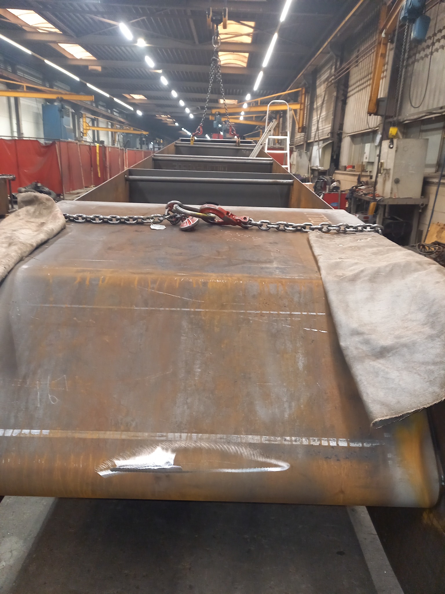 Metalowa konstrukcja z łańcuchami i hakami zawieszona pod sufitem hali produkcyjnej, widoczne ślady spawania i obróbki.