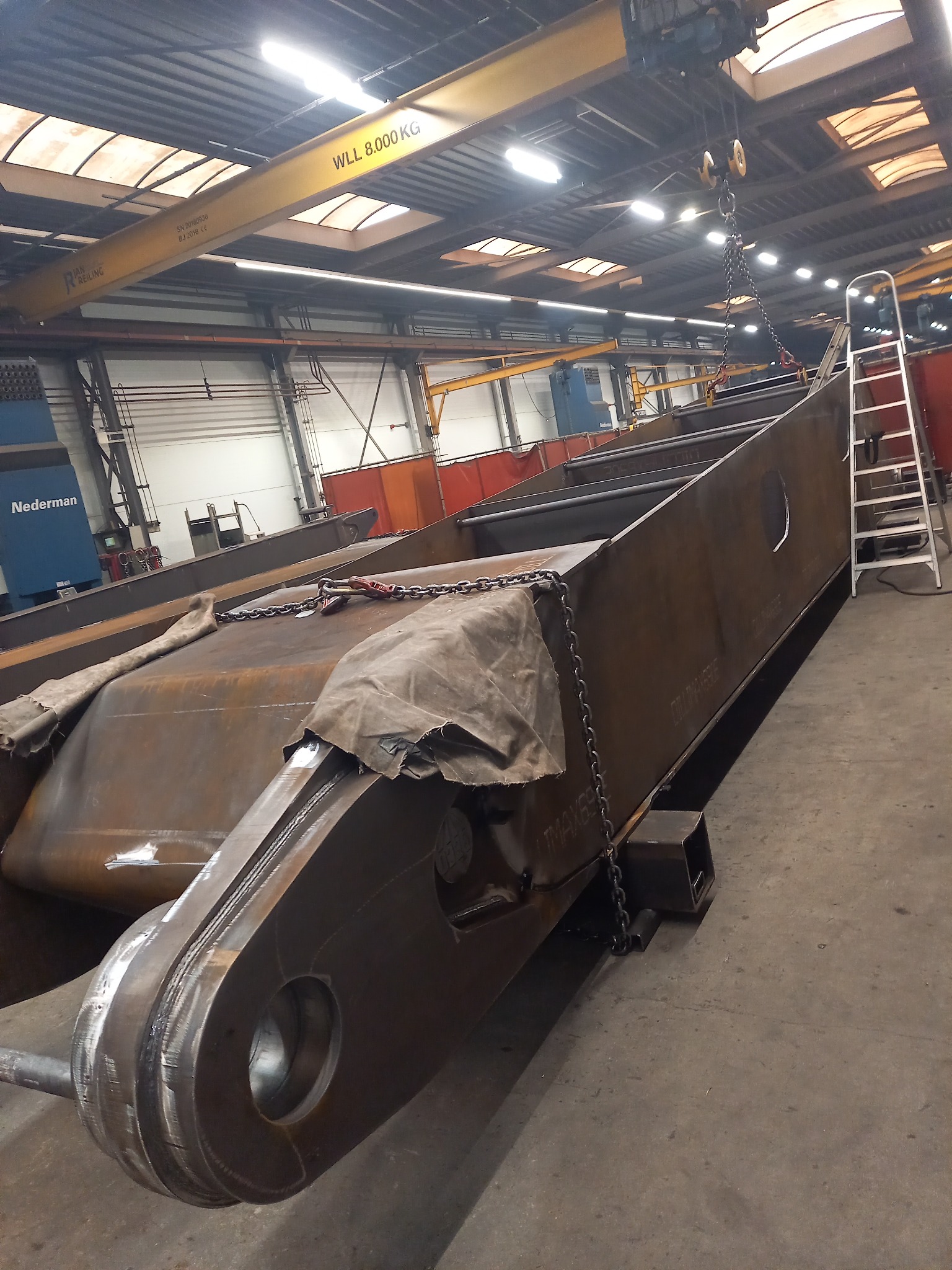 Fragment konstrukcji stalowej z widocznymi spawami, podnoszonej za pomocą łańcucha przez suwnicę w hali produkcyjnej w Osjakowie. Widoczna drabina i maszyna do cięcia plazmowego.