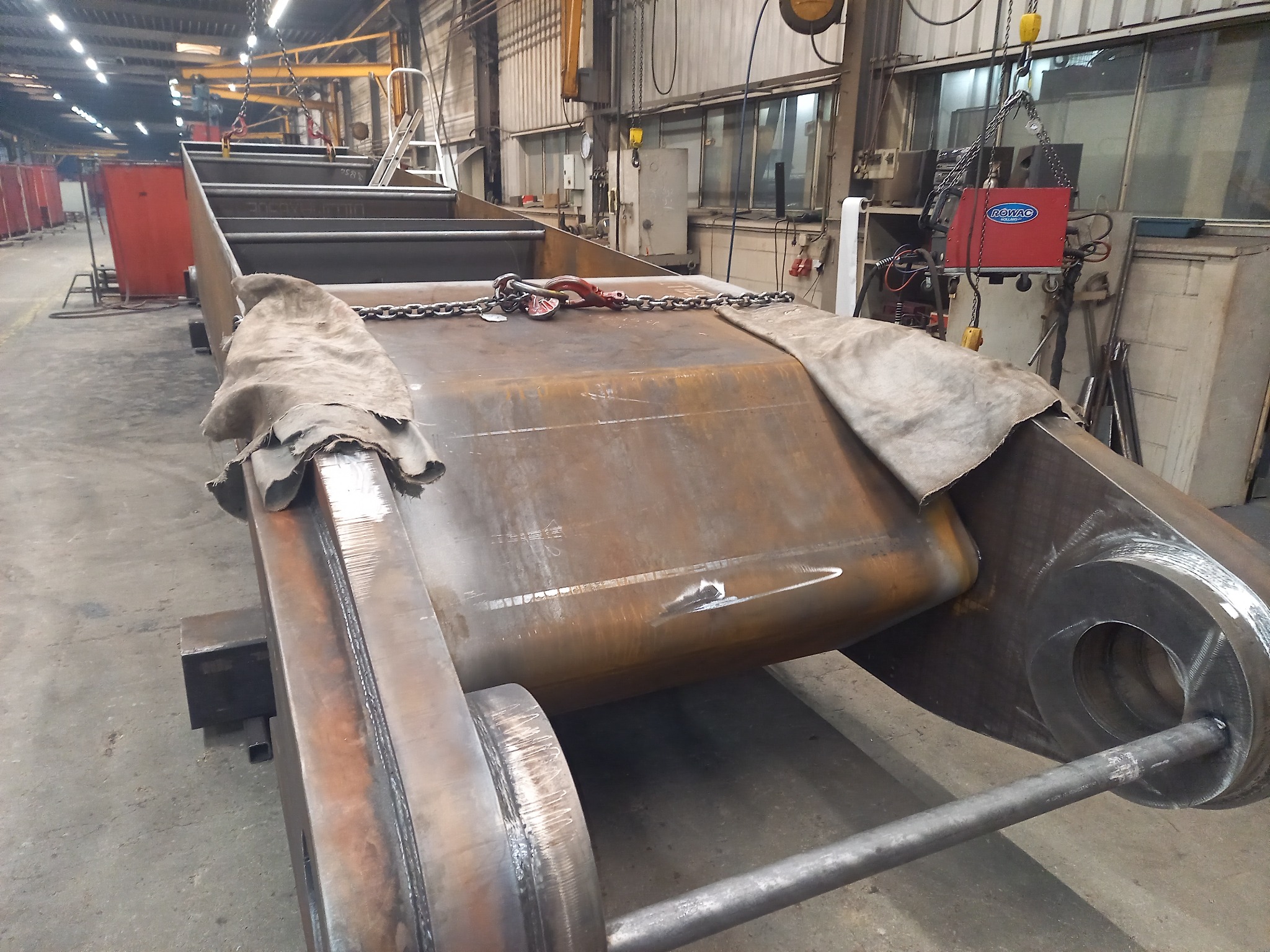 Część konstrukcji metalowej w hali produkcyjnej, zabezpieczona płótnem, z widocznymi spawami i elementami mocującymi, w tle maszyna spawalnicza i drabina.