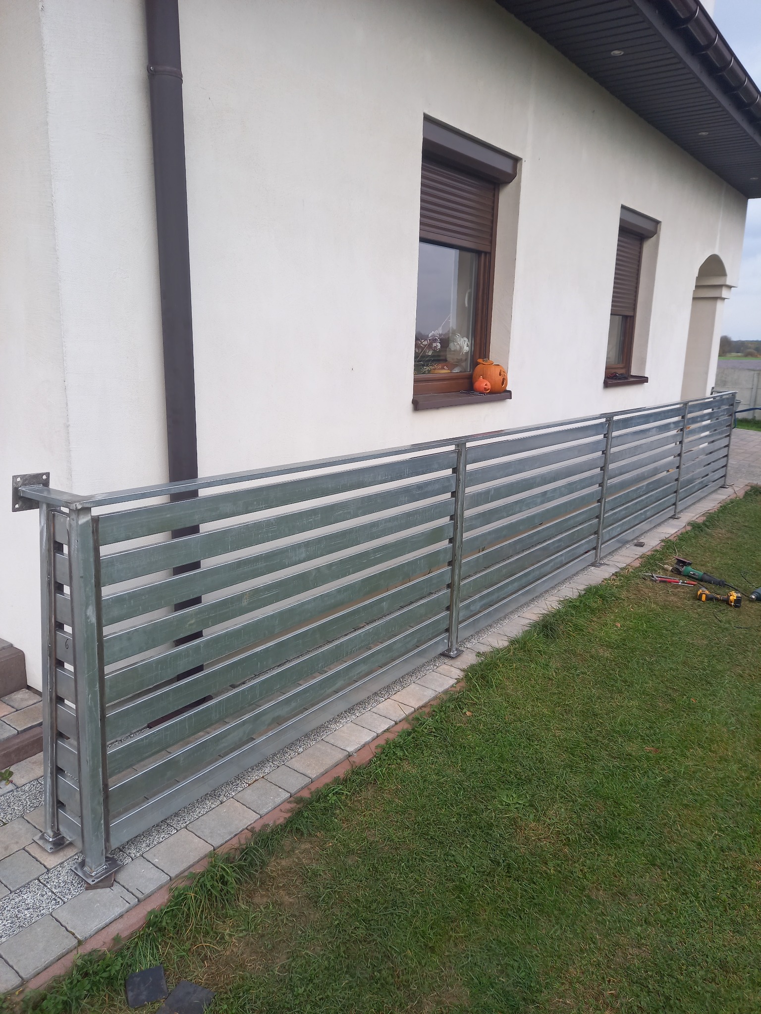 Nowoczesne ogrodzenie metalowe z poziomych profili, surowy metal, widoczne narzędzia na trawie, dom z brązowymi roletami w tle.