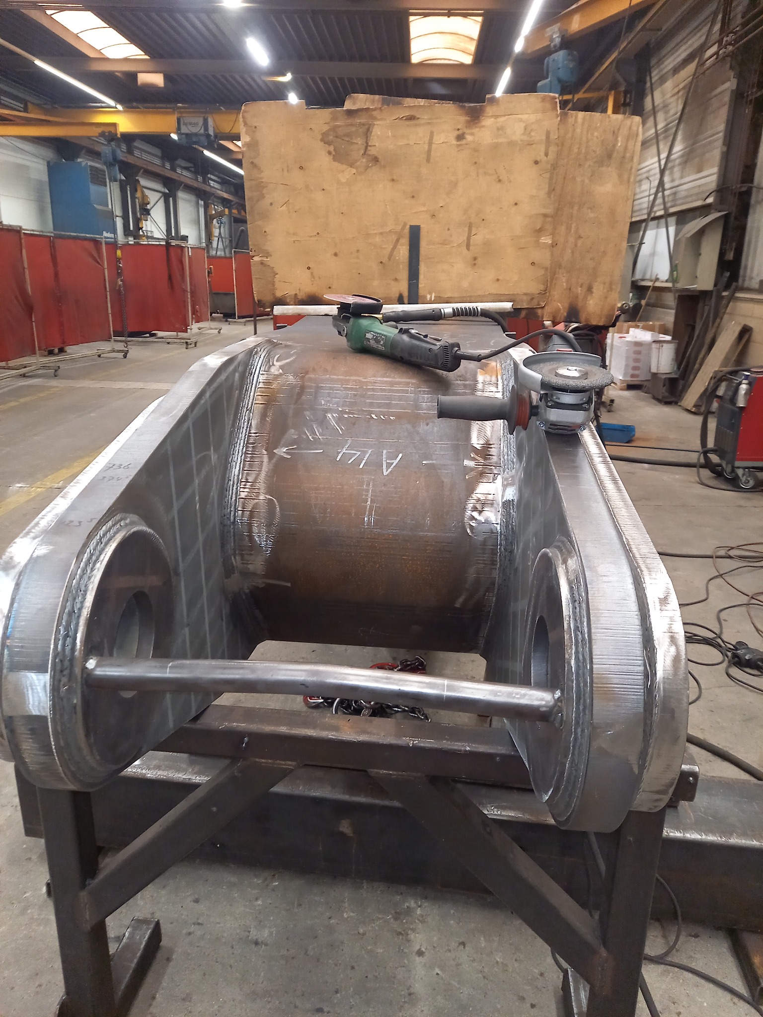 Duży, spawany stalowy element konstrukcyjny na metalowym stojaku w hali produkcyjnej, obok szlifierki kątowe i spawarka.