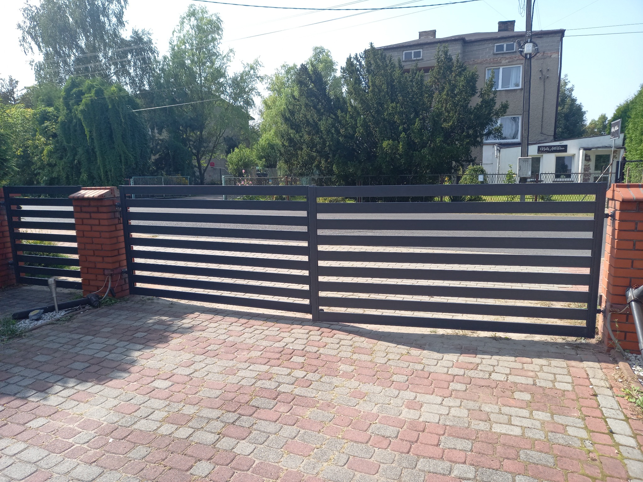 Dwuskrzydłowa brama wjazdowa z poziomych profili, osadzona na ceglanych słupkach, widoczna z zewnątrz posesji na tle budynku i zieleni.