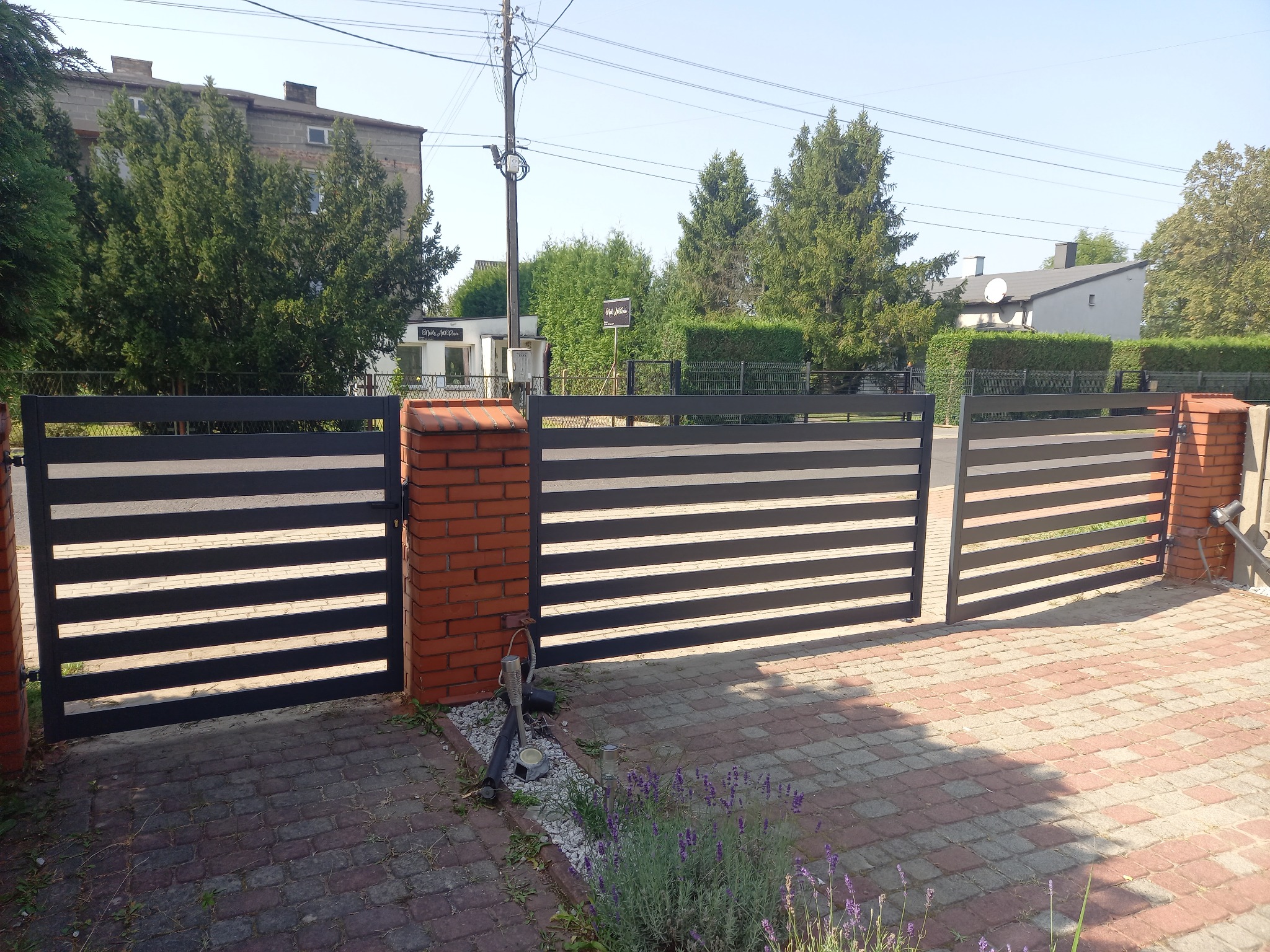 Nowoczesna brama wjazdowa z poziomymi, czarnymi przęsłami, osadzona na ceglanych słupkach, z widoczną kostką brukową i zielenią w tle.