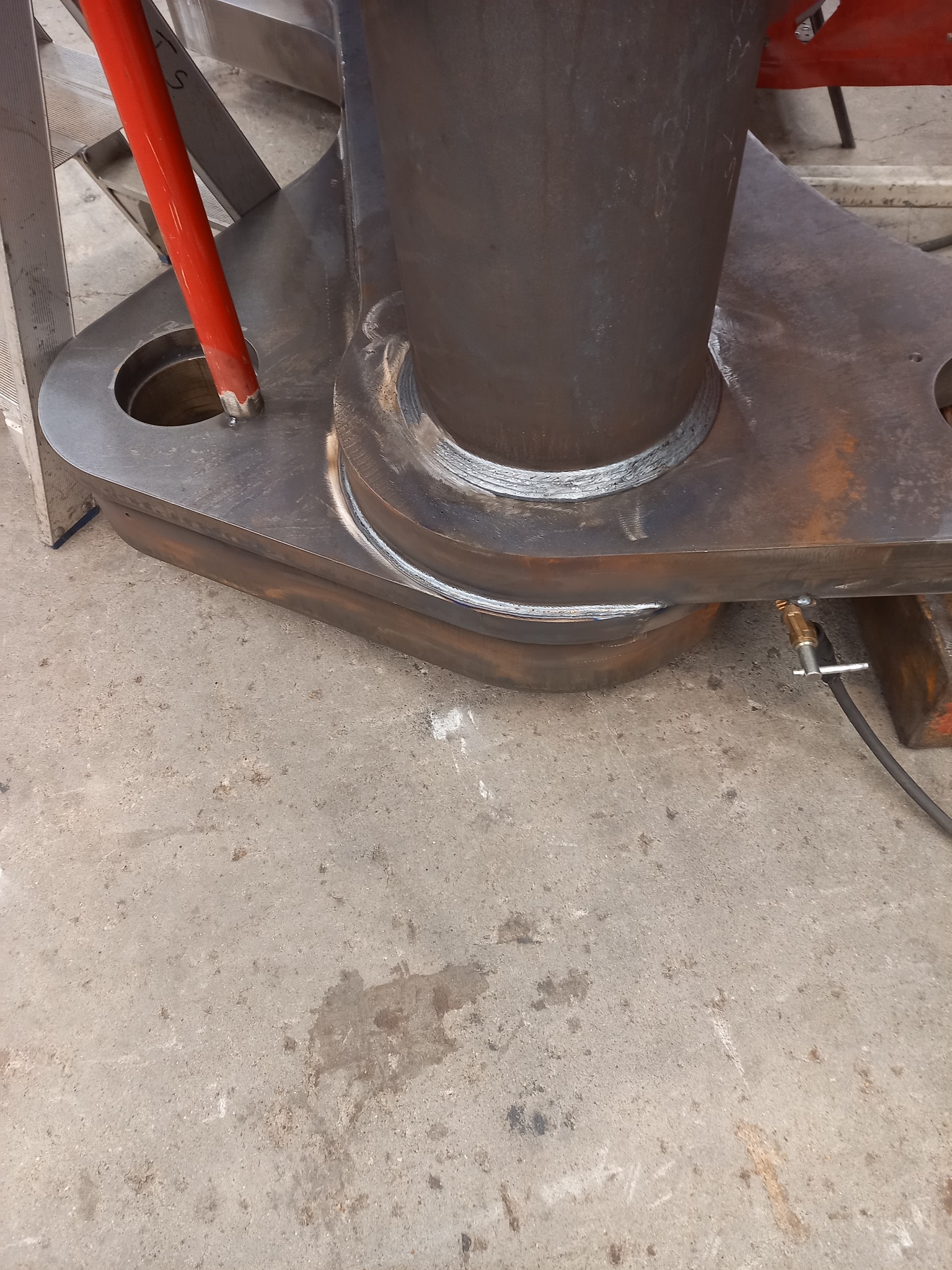 Metalowy element konstrukcyjny po spawaniu, widoczne spoiny, rura wtopiona w grubą płytę z otworami, szary beton na podłodze.