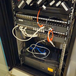 ENERGY - TELETECH CEZARY WIŚNIEWSKI - Szafa rack z zainstalowanymi urządzeniami sieciowymi: switche, router, zasilacze z etykietami Orange, CCTV, Drytec, okablowanie w kolorach białym, niebieskim i pomarańczowym.