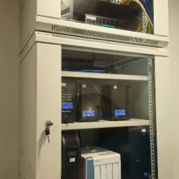 Dwie szafy rack zamontowane jedna nad drugą, z widocznym okablowaniem, przełącznikami sieciowymi, zasilaczami UPS i serwerami NAS, zabezpieczone zamkami na klucz.