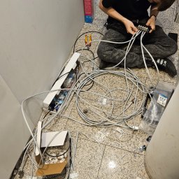 ENERGY - TELETECH CEZARY WIŚNIEWSKI - Młody instalator w czarnej koszulce i dżinsach klęczy na podłodze z terakoty, podłączając kable do rozdzielacza; obok leżą rozpakowane kable, narzędzia i pudełka z elementami montażowymi.