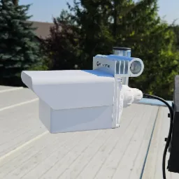 Biała antena Mikrotik na dachu z widocznym kablem, zamontowana na tle zielonych drzew i błękitnego nieba. Widok z bliska, podkreślający detale urządzenia.