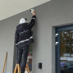 ENERGY - TELETECH CEZARY WIŚNIEWSKI - Instalator na drabinie montuje kamerę monitoringu na elewacji budynku. Widoczna boczna ściana budynku z oknem i fragmentem zaparkowanego samochodu.