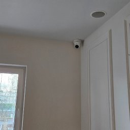 ENERGY - TELETECH - Kamera monitoringu zamontowana pod sufitem w pomieszczeniu z drabiną. Widoczny fragment okna i ozdobna ściana. Ujęcie z dołu.