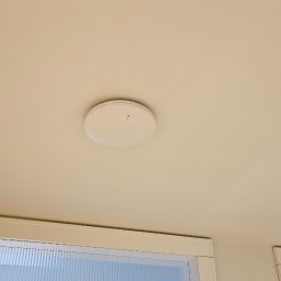 ENERGY - TELETECH CEZARY WIŚNIEWSKI - Zamontowany, okrągły czujnik dymu na białym suficie w pomieszczeniu z oknem. Widoczny fragment drewnianej zabudowy. Ujęcie z dołu, centralnie.