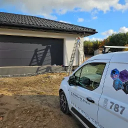 Drabina oparta o białą ścianę budynku z zamontowaną czarną bramą garażową, obok zaparkowany biały samochód firmowy z logo i numerem telefonu, widok na błękitne niebo z chmurami.