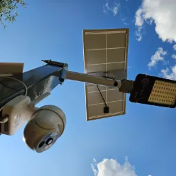 Kamera monitoringu PTZ oraz panel solarny i lampa LED zamontowane na słupie, widok z dołu na tle błękitnego nieba z chmurami