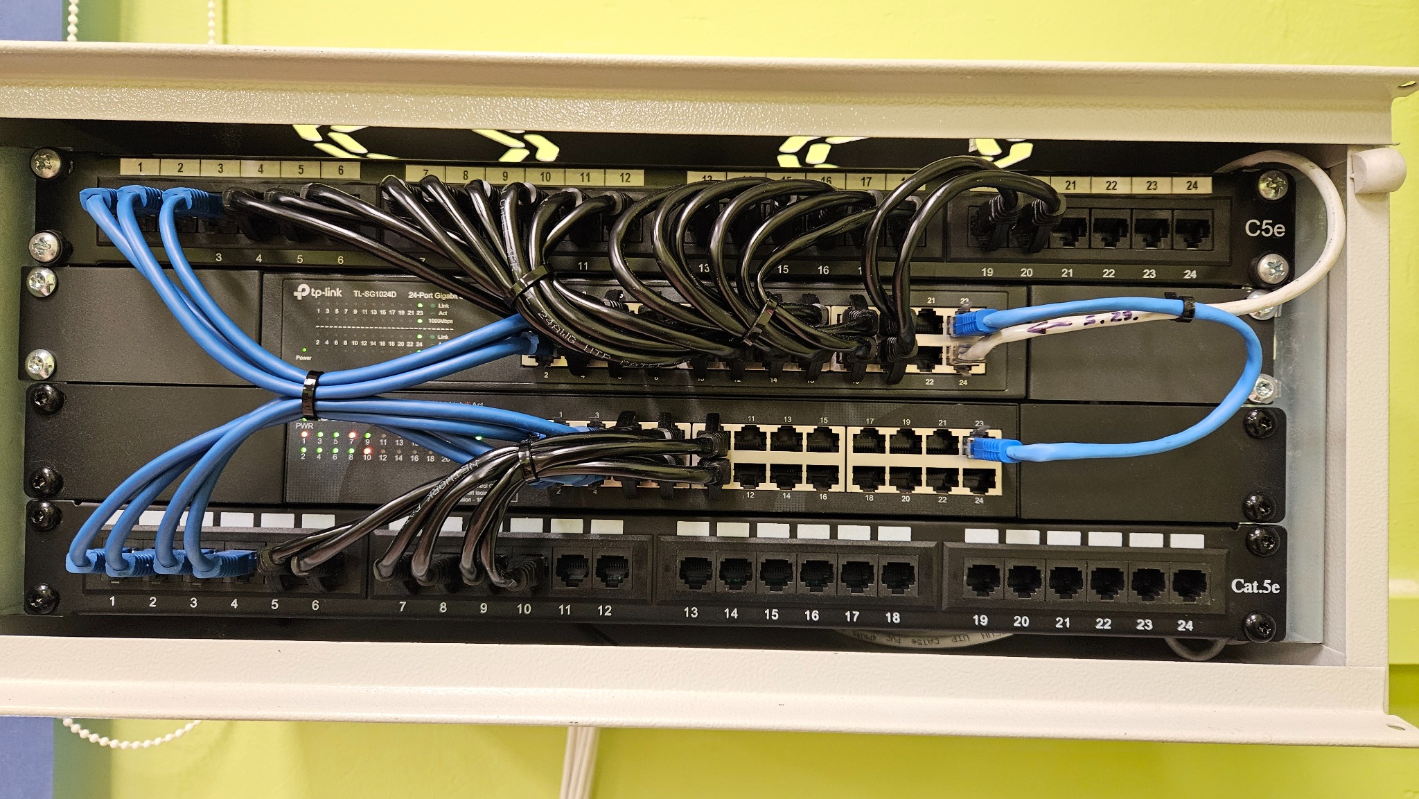 Panel krosowniczy z podłączonymi kablami sieciowymi Ethernet, część kabli spięta opaskami zaciskowymi, widoczny switch TP-Link TL-SG1024D, oznaczenia portów C5e i Cat.5e.