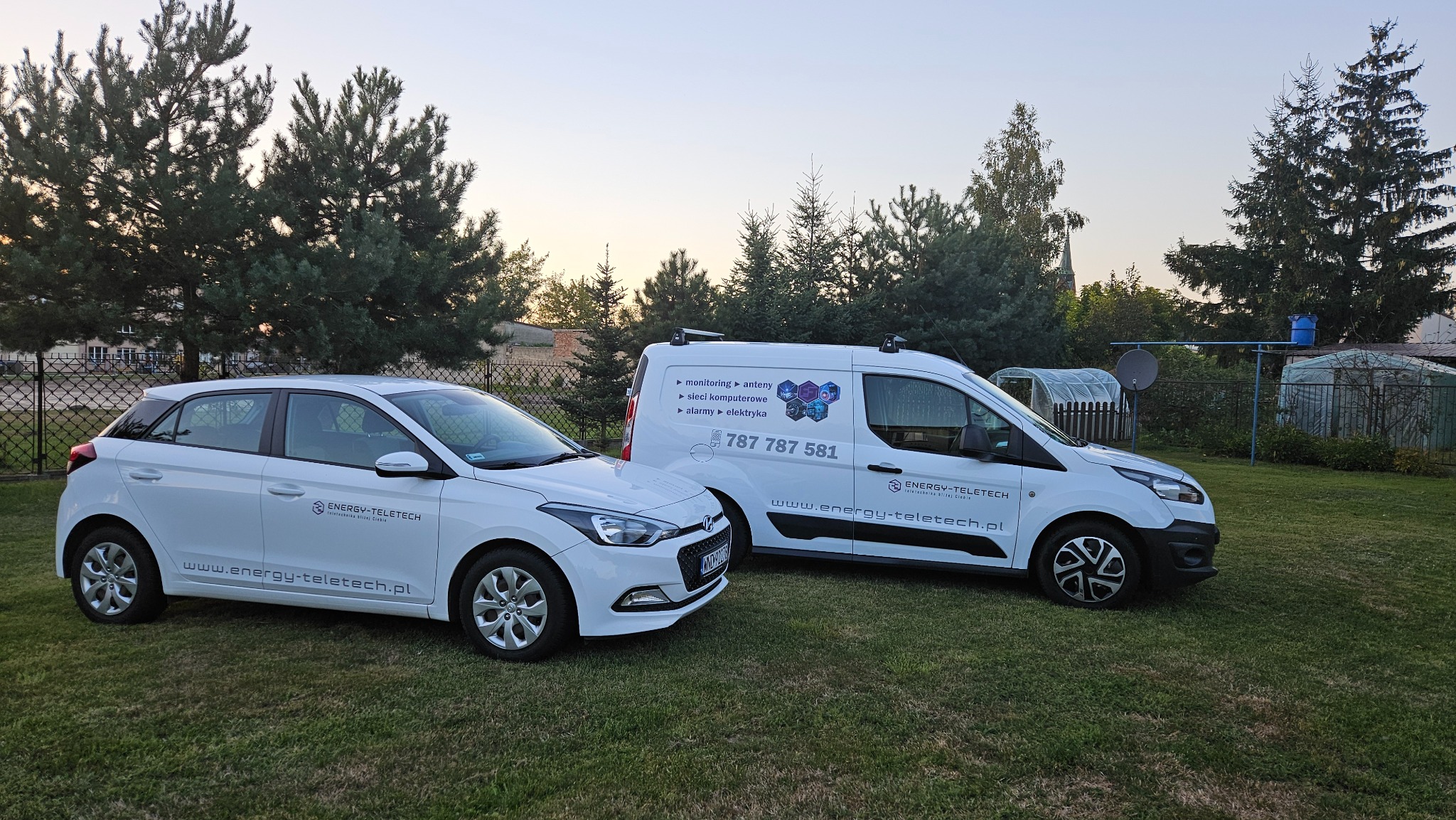 Dwa białe samochody firmy Energy-Teletech z reklamowymi napisami i logo na tle zielonego trawnika i drzew iglastych.