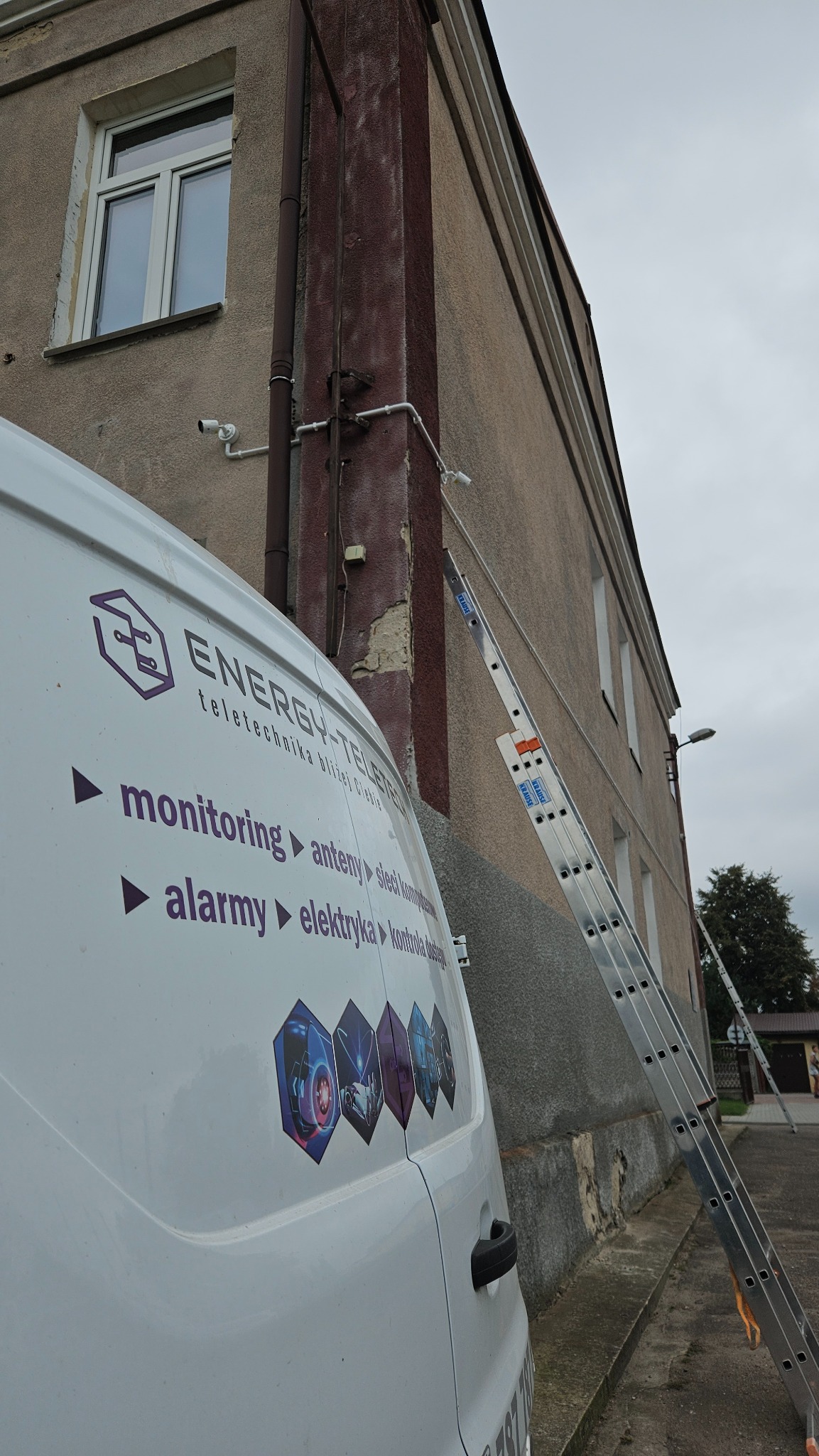 Kamera monitoringu zamontowana na elewacji budynku, widoczna drabina oparta o ścianę oraz samochód dostawczy z logo firmy telekomunikacyjnej.