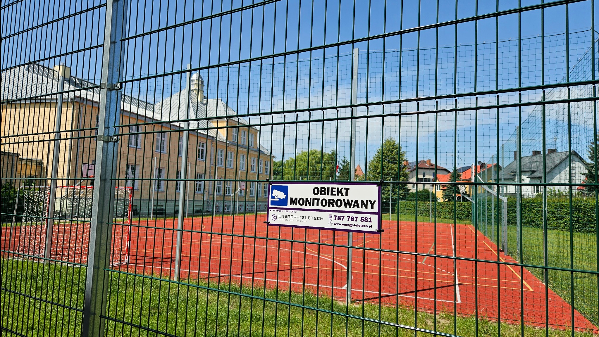 Tablica informacyjna 'Obiekt Monitorowany' umieszczona na ogrodzeniu boiska szkolnego z bramkami, w tle budynek szkoły i domy jednorodzinne