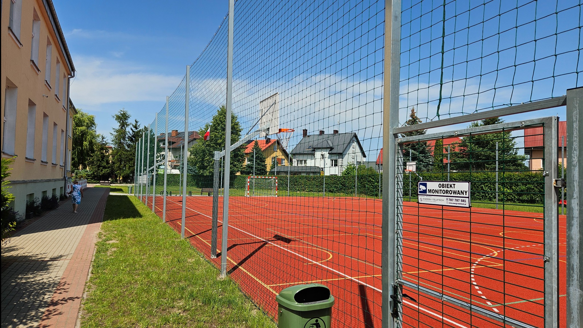 Ogrodzone boisko sportowe z czerwoną nawierzchnią, widoczne bramki do piłki nożnej i kosz do koszykówki, tabliczka informująca o monitoringu, zieleń w tle, błękitne niebo.