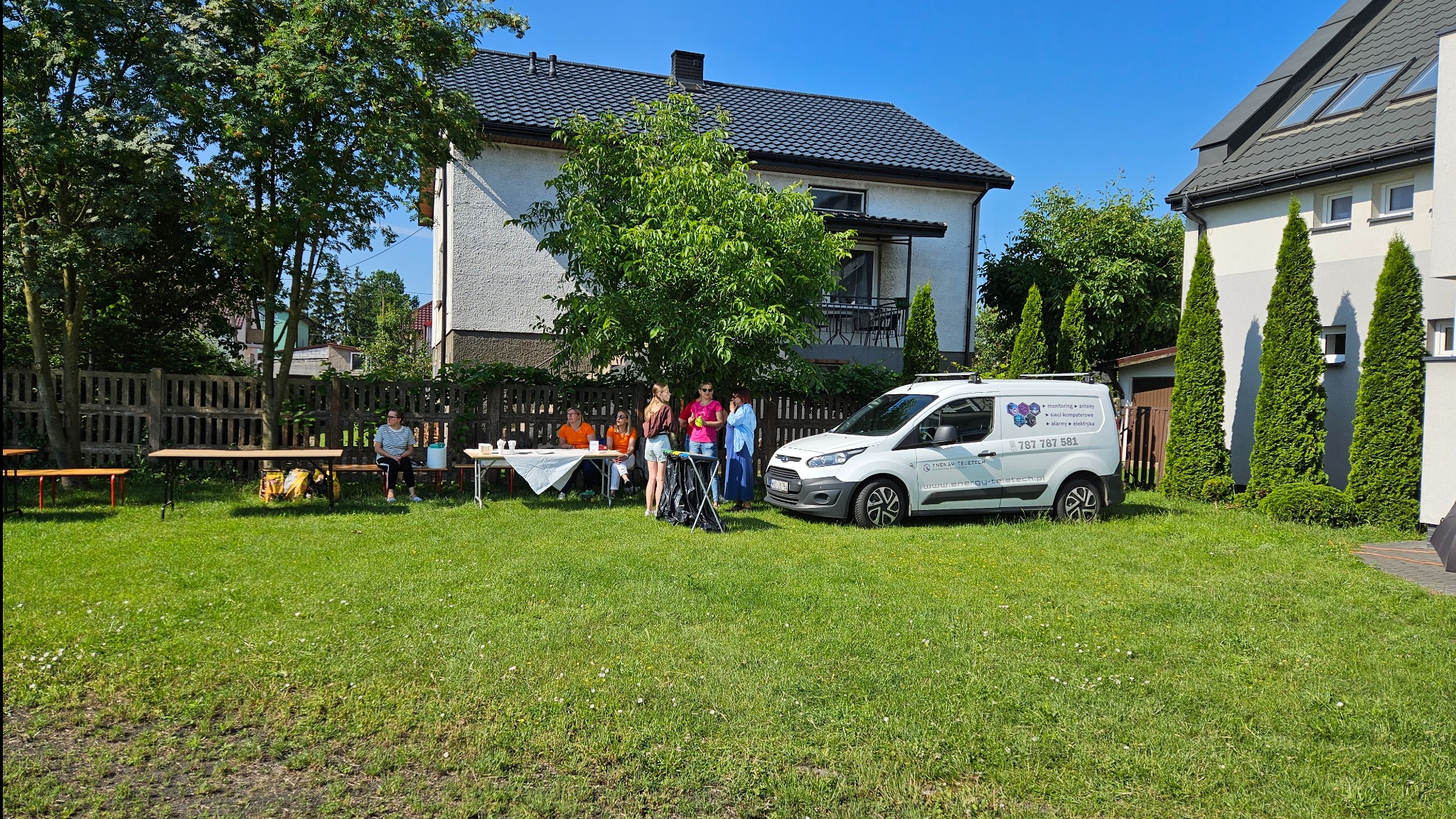 Biały samochód dostawczy z logo firmy telekomunikacyjnej zaparkowany na trawniku przed domem jednorodzinnym, obok grupa osób przy stole.
