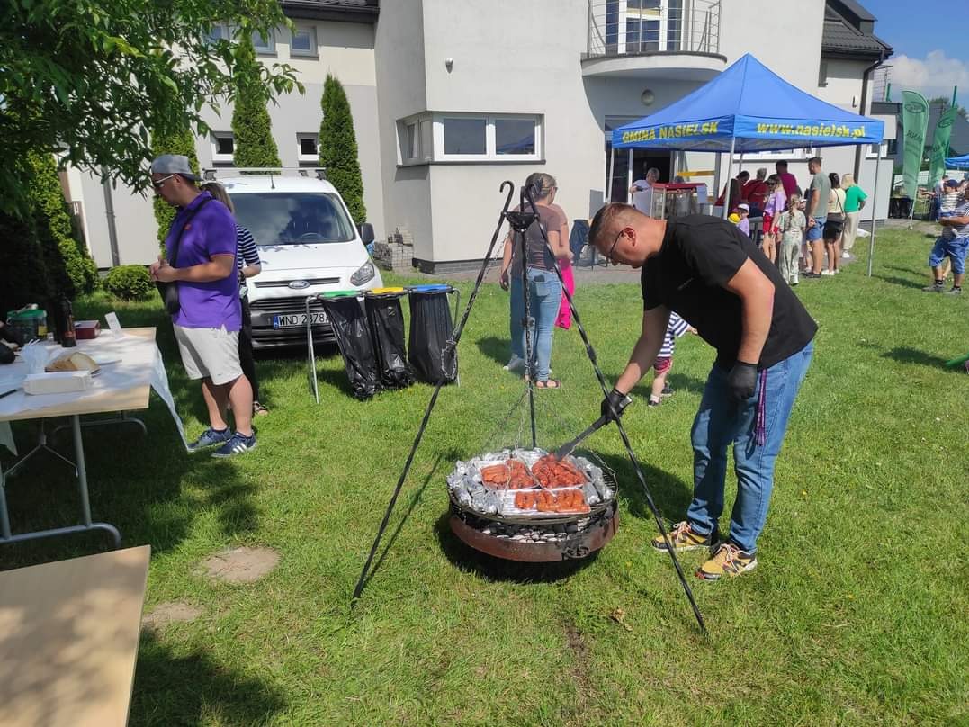 Mężczyzna w czarnej koszulce i jeansach grilluje mięso na trójnogu nad paleniskiem z węglem drzewnym, na trawie, w tle budynek, biały van i grupa osób.