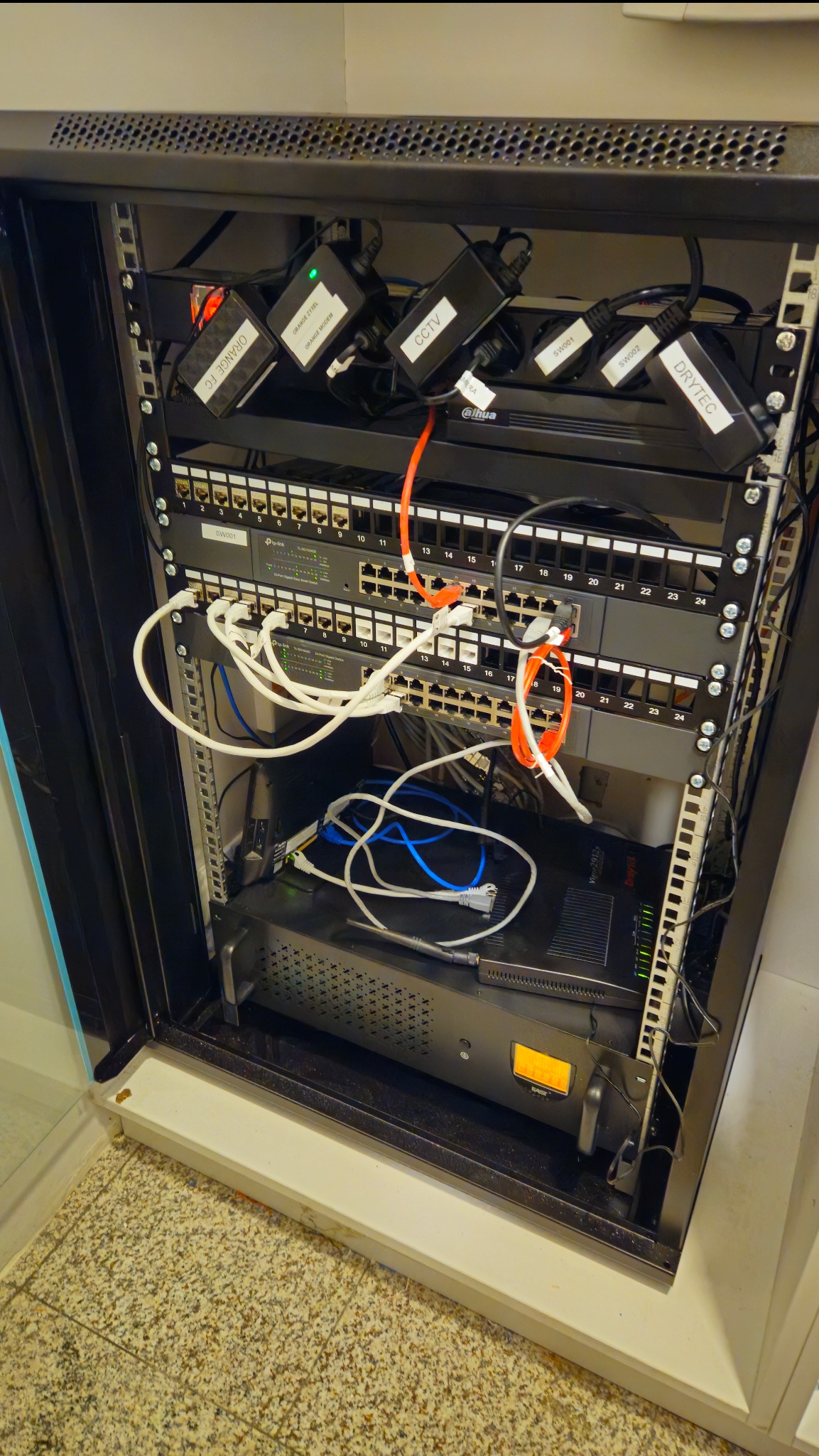 Szafa rack z zainstalowanymi urządzeniami sieciowymi: switche, router, zasilacze z etykietami Orange, CCTV, Drytec, okablowanie w kolorach białym, niebieskim i pomarańczowym.