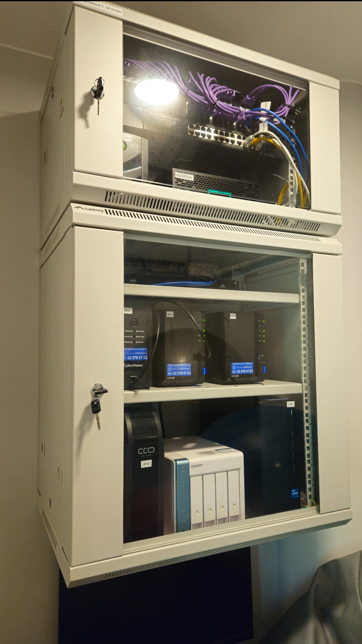 Dwie szafy rack zamontowane jedna nad drugą, z widocznym okablowaniem, przełącznikami sieciowymi, zasilaczami UPS i serwerami NAS, zabezpieczone zamkami na klucz.