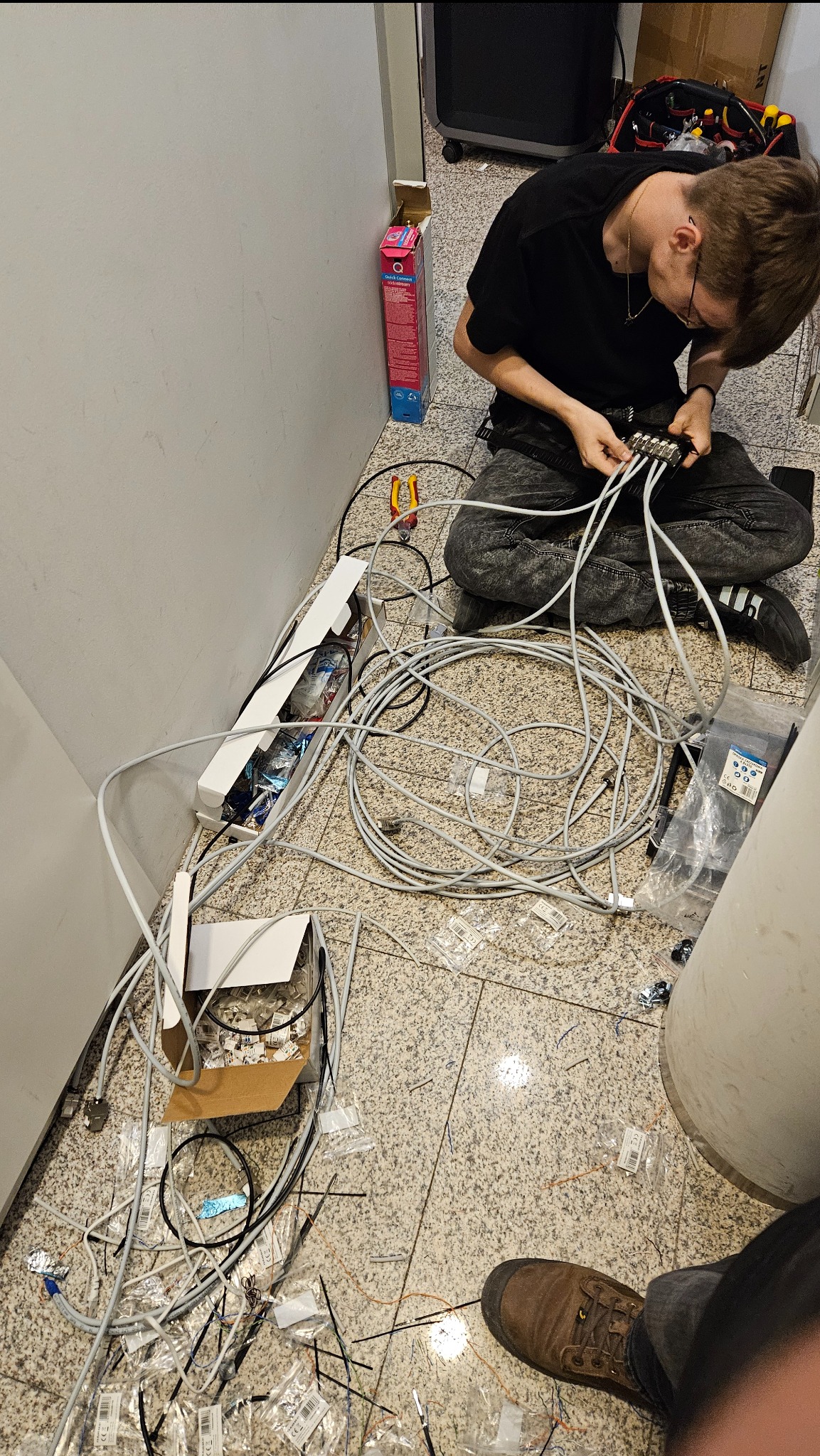 Młody instalator w czarnej koszulce i dżinsach klęczy na podłodze z terakoty, podłączając kable do rozdzielacza; obok leżą rozpakowane kable, narzędzia i pudełka z elementami montażowymi.