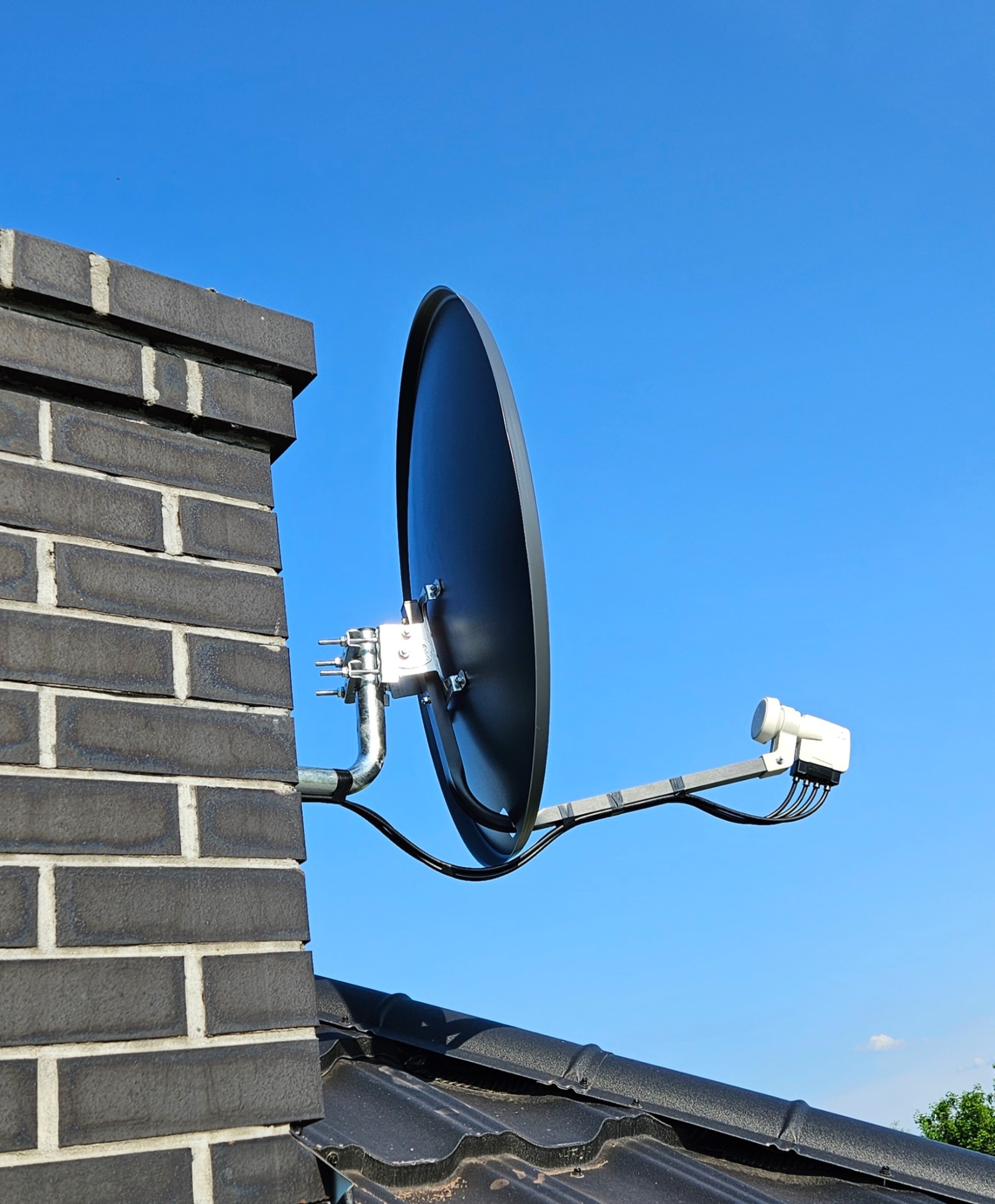 Czarna antena satelitarna zamontowana na kominie z ciemnej cegły na tle błękitnego nieba, widok z dołu.