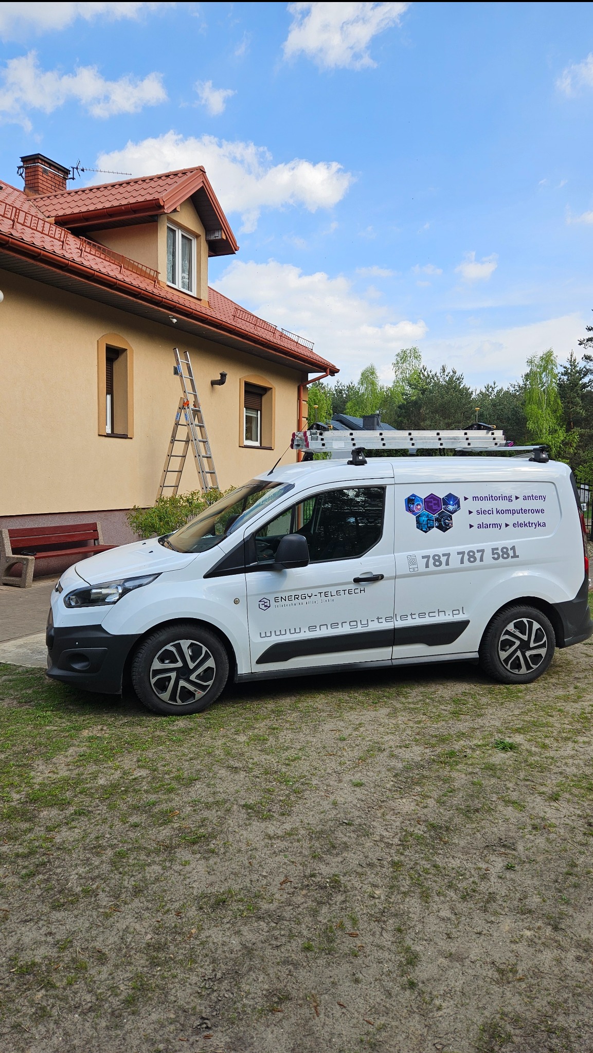 Biały samochód dostawczy firmy Energy-Teletech z drabiną na dachu zaparkowany przed domem z czerwoną dachówką; na samochodzie widoczne logo i zakres usług: monitoring, anteny, sieci komputerowe...