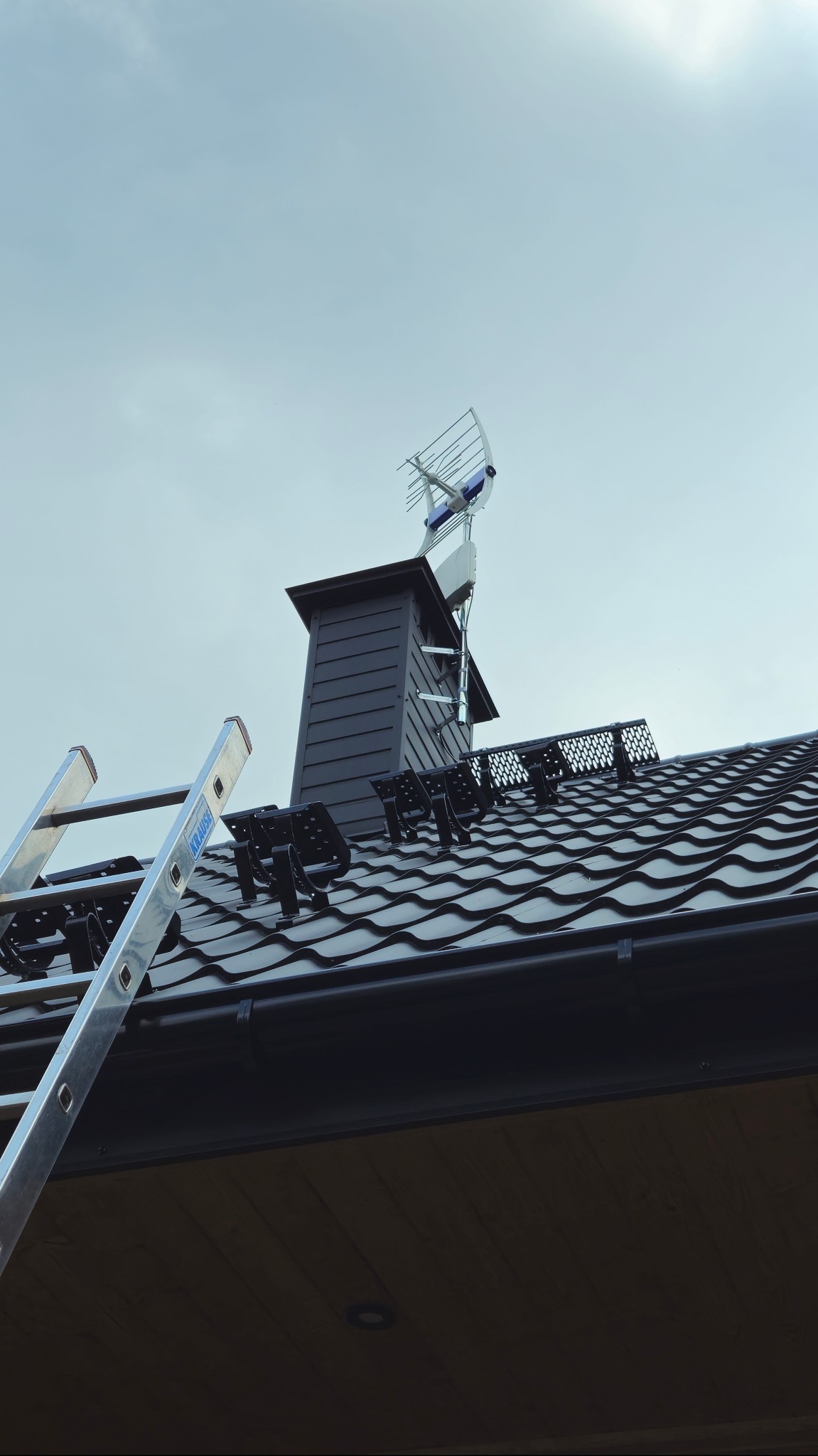Antena telewizyjna na dachu z metalową dachówką, widoczna drabina Krause. Ujęcie od dołu, błękitne niebo w tle.