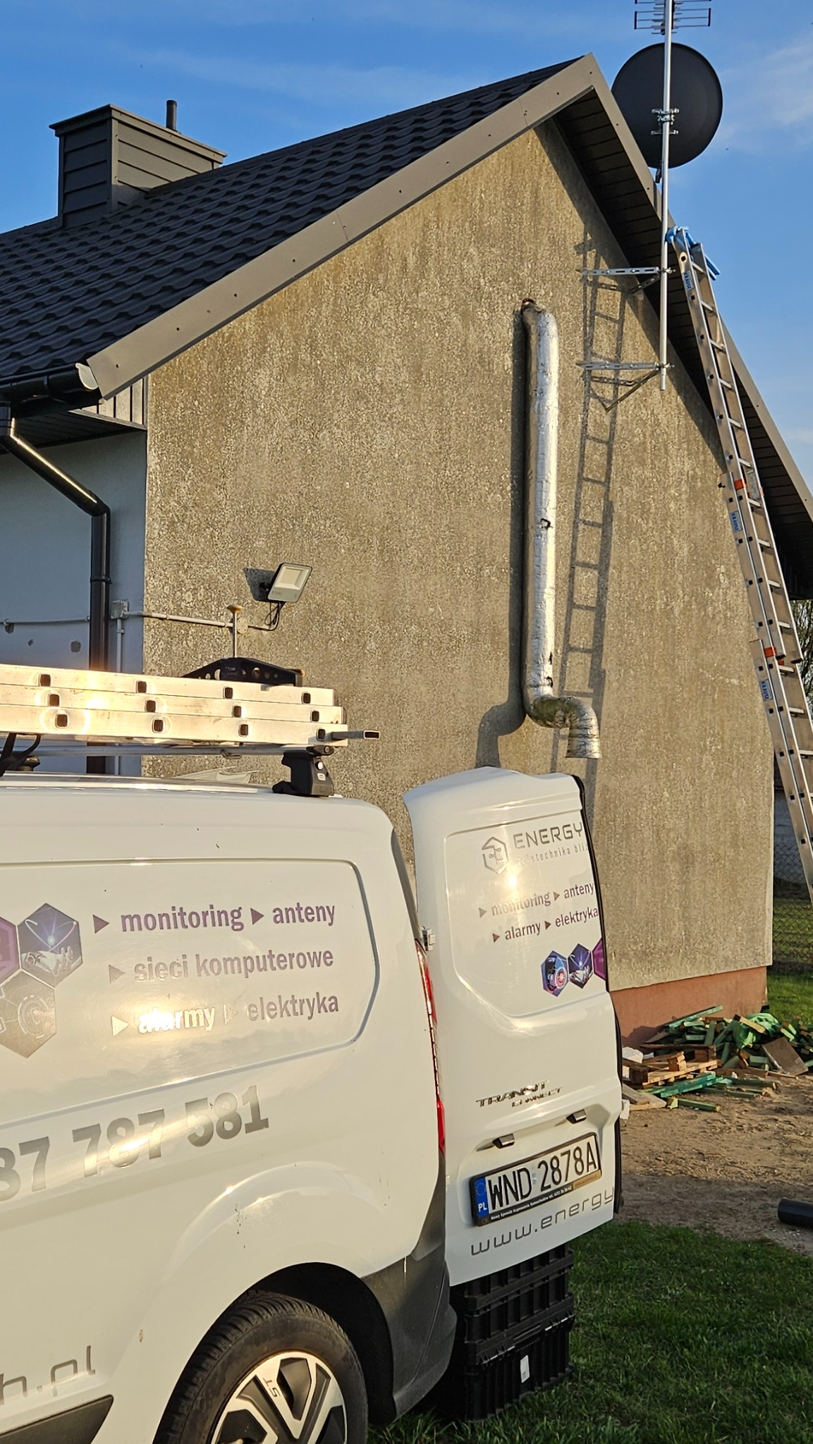 Antena satelitarna na dachu domu, drabina oparta o ścianę, samochód z logo firmy instalacyjnej. Usługi: monitoring, anteny, sieci, alarmy, elektryka.