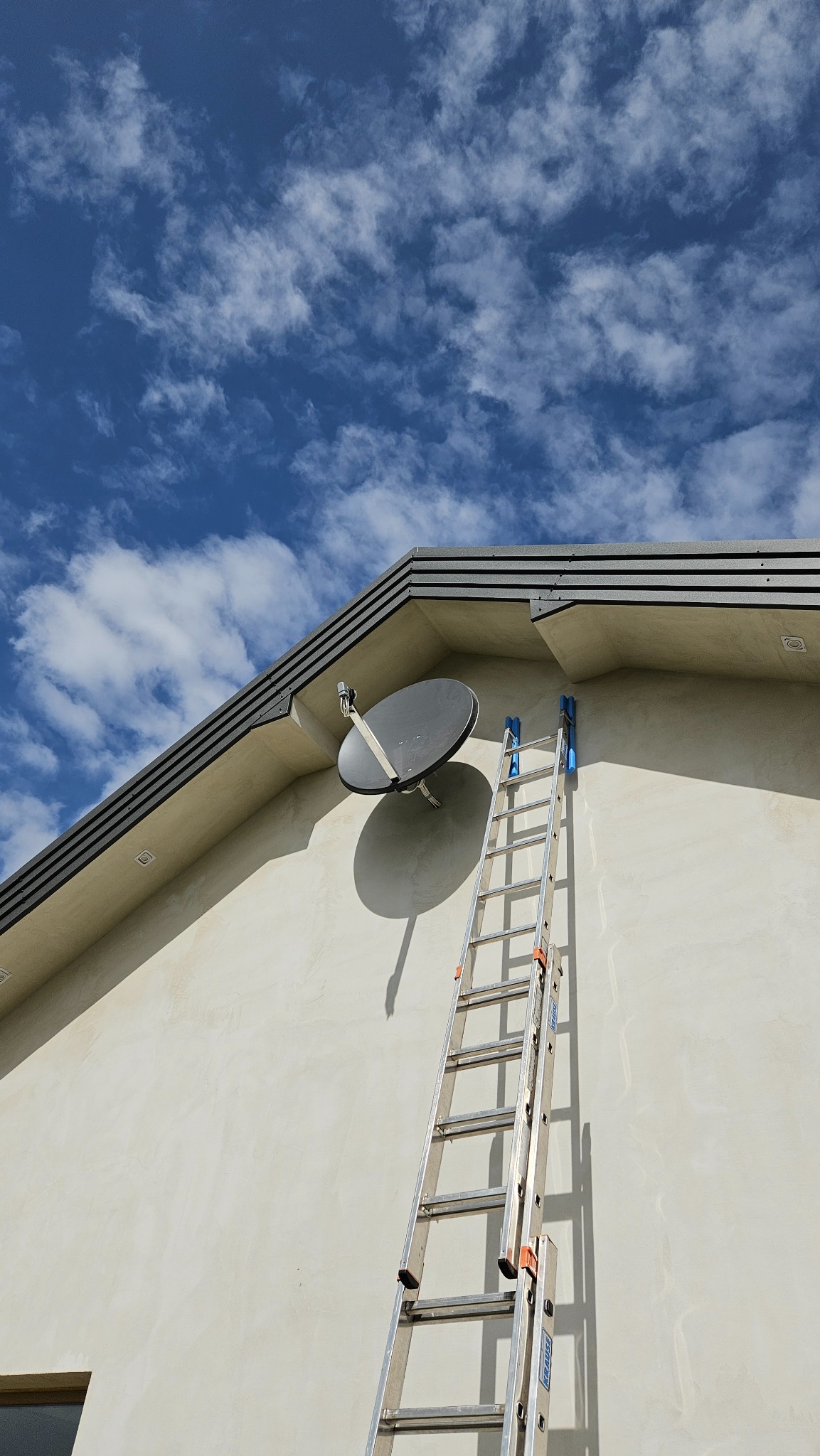 Antena satelitarna zamontowana na elewacji domu, z drabiną opartą o ścianę. Ujęcie z dołu, widoczne błękitne niebo z chmurami. Nowoczesne wykończenie budynku.