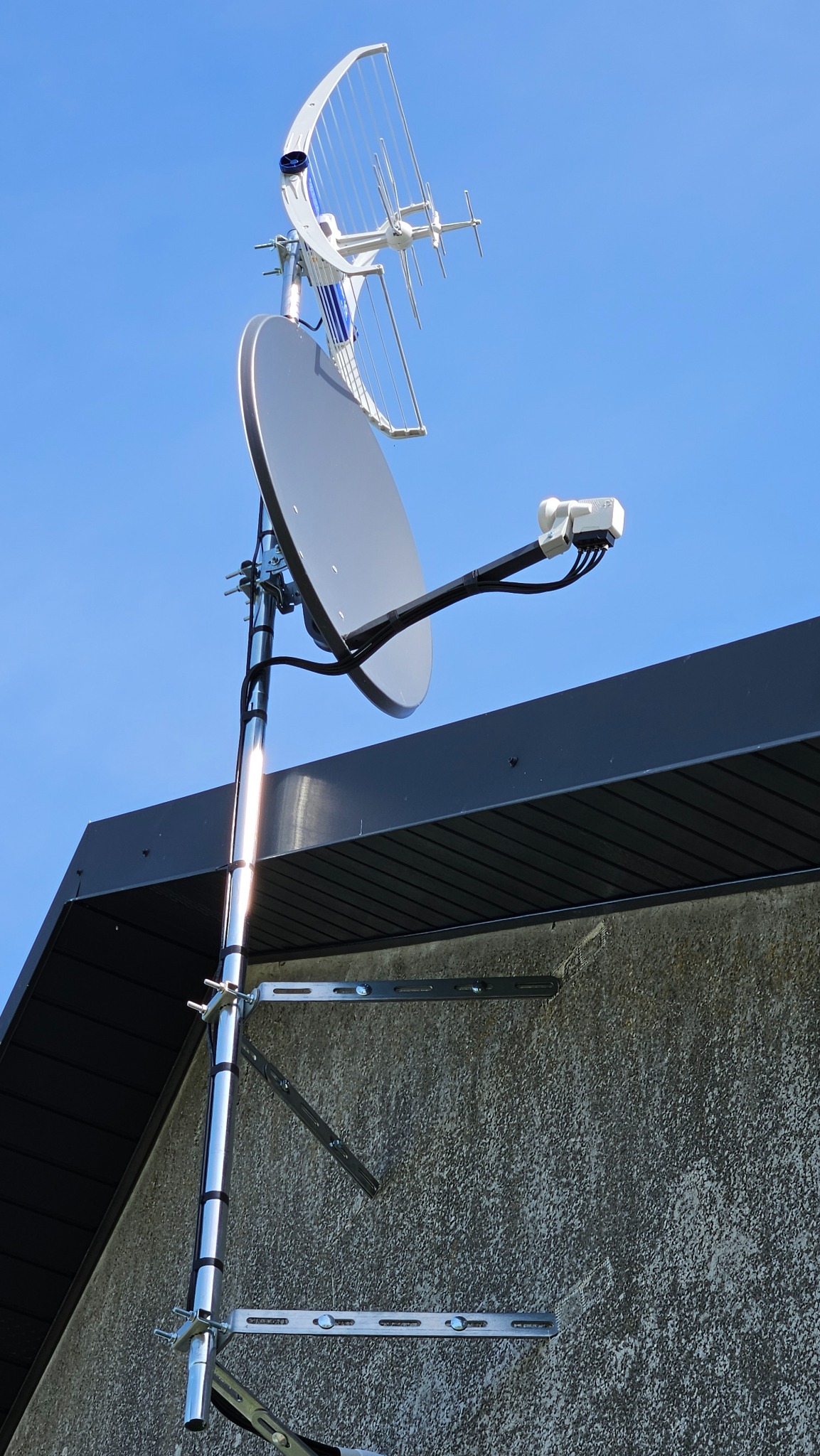 Antena satelitarna i antena kierunkowa VHF/UHF zamontowane na dachu budynku, z widocznymi elementami mocującymi i okablowaniem. Jasne, błękitne niebo w tle.