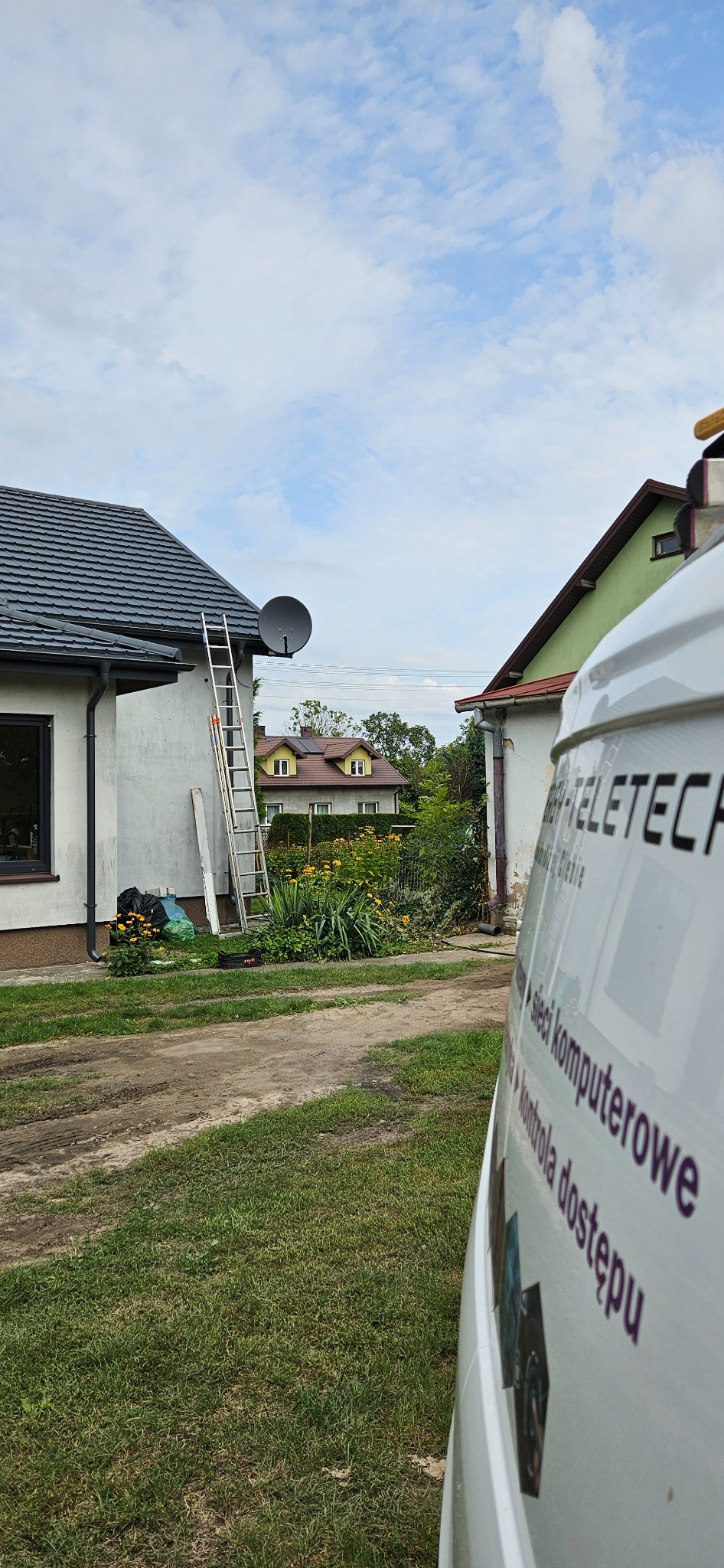 Antena satelitarna zamontowana na ścianie domu, drabina oparta o ścianę, widoczny fragment firmowego auta z napisem 'sieci komputerowe'.