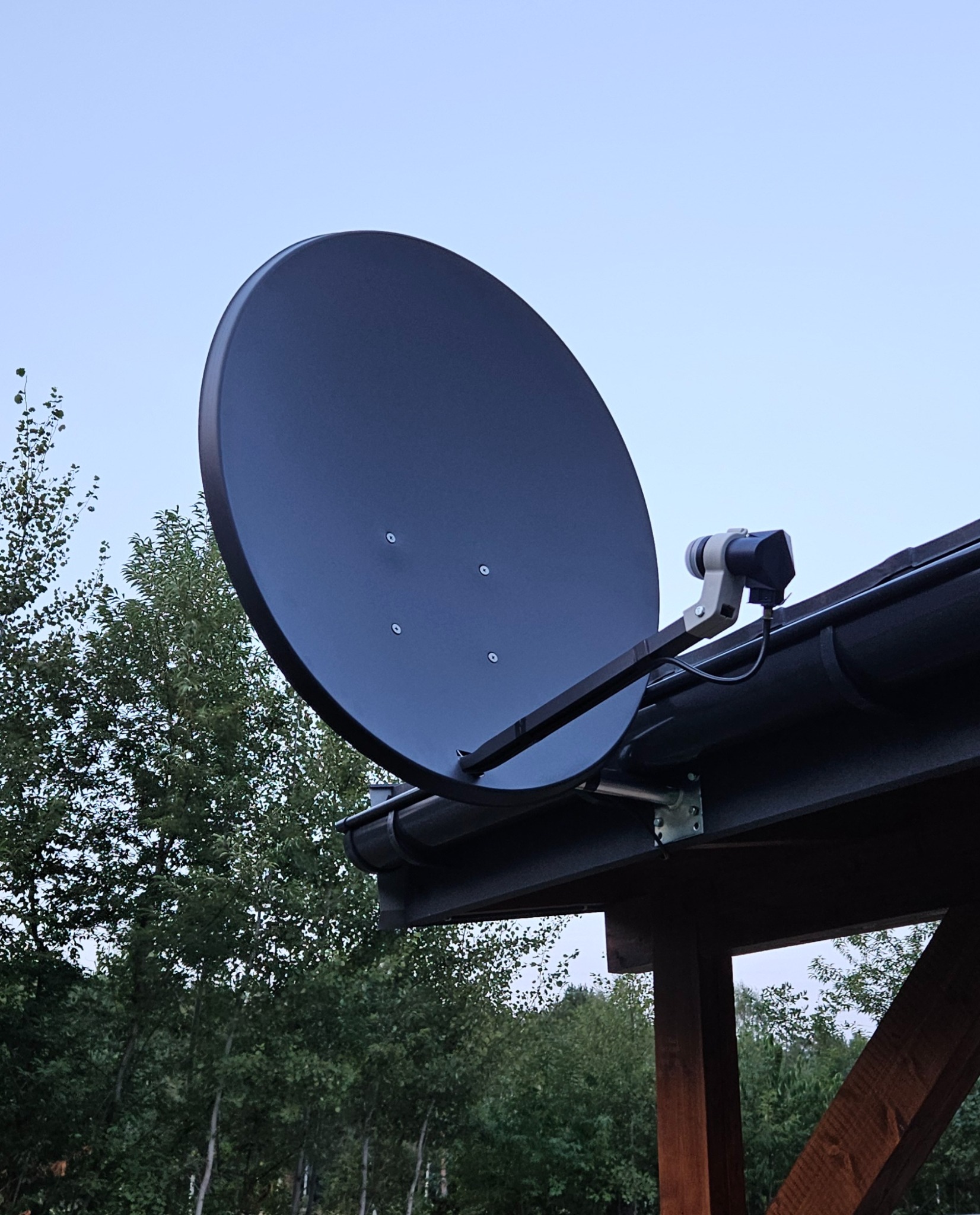 Czarna antena satelitarna zamontowana na dachu budynku z widocznymi drzewami w tle. Ujęcie z dołu, podkreślające instalację antenową.