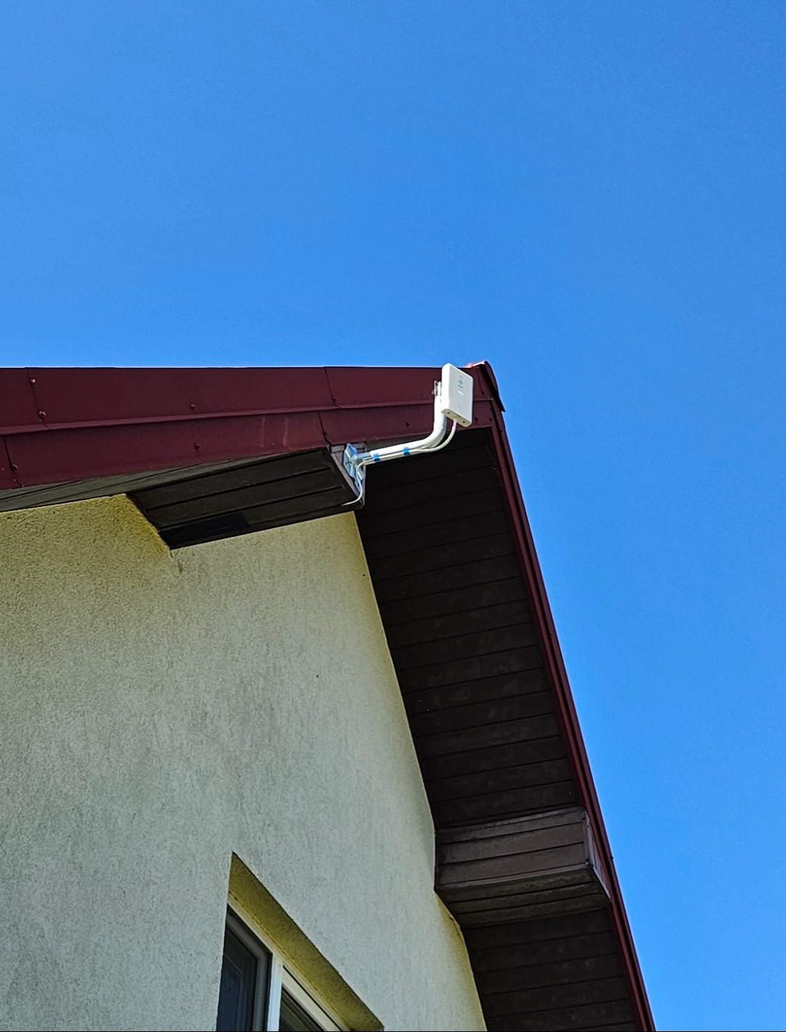 Biała skrzynka z okablowaniem zamontowana na rogu dachu domu z czerwoną dachówką, na tle błękitnego nieba. Widok z dołu.