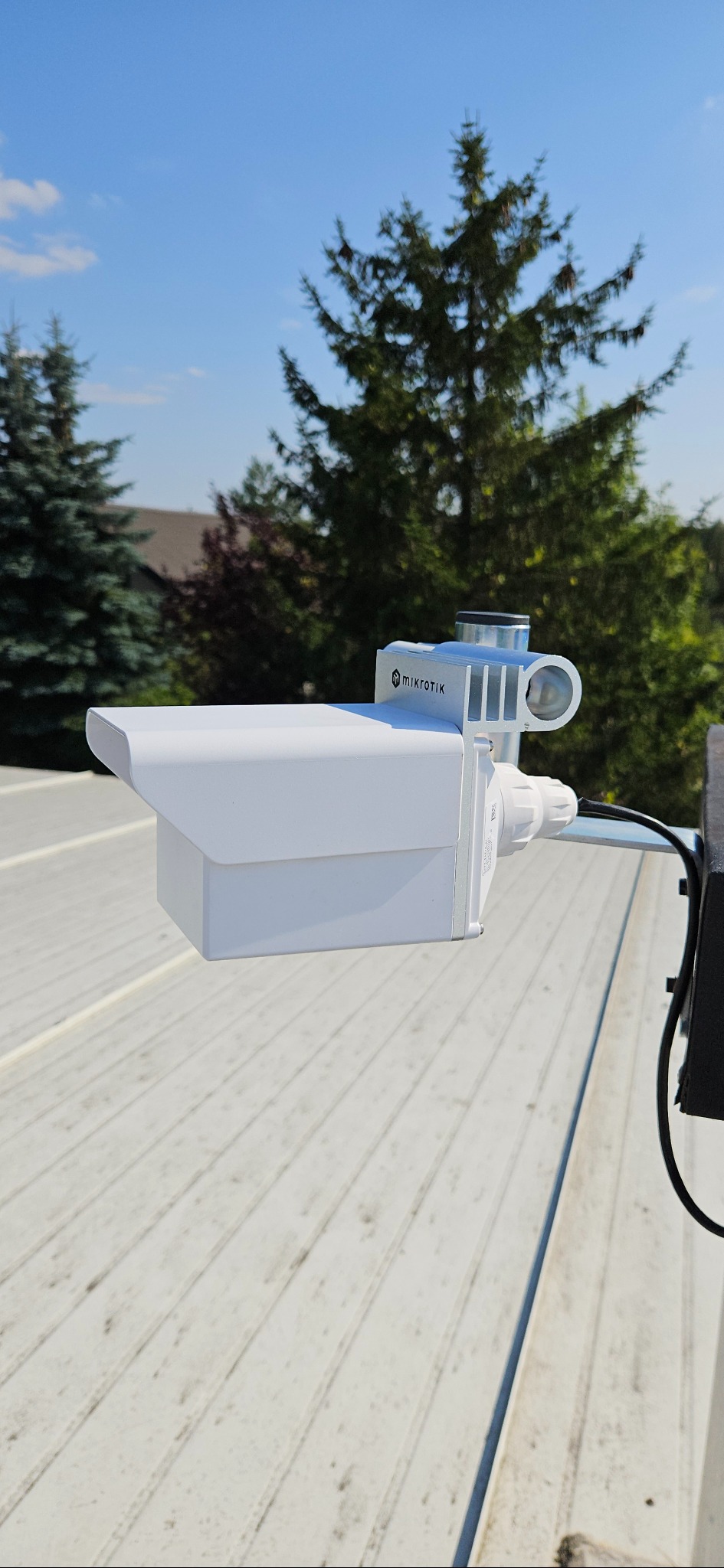 Biała antena Mikrotik na dachu z widocznym kablem, zamontowana na tle zielonych drzew i błękitnego nieba. Widok z bliska, podkreślający detale urządzenia.