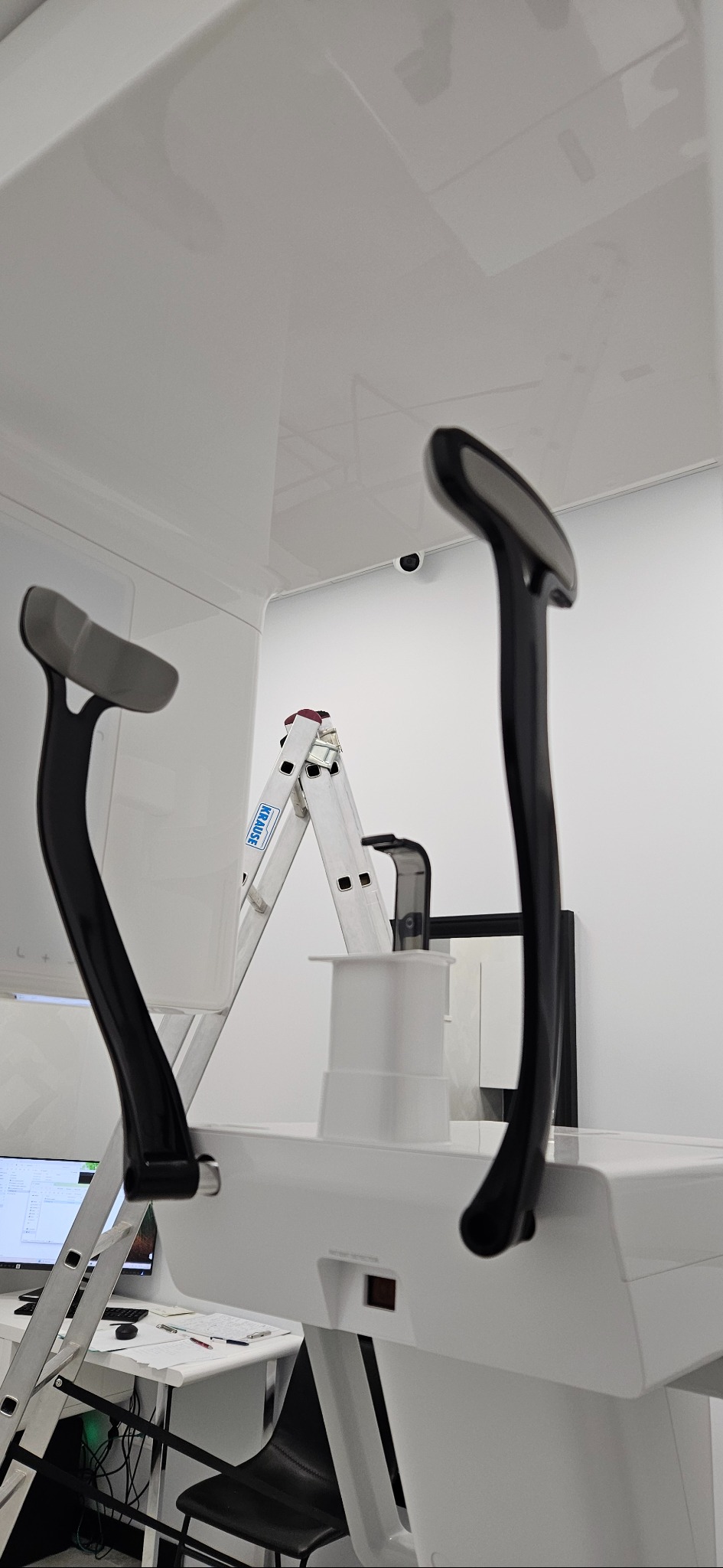 Wnętrze z białą drabiną Krause, nowoczesnym urządzeniem diagnostycznym i zamontowaną kamerą monitoringu na suficie. Minimalistyczny design, jasne oświetlenie.