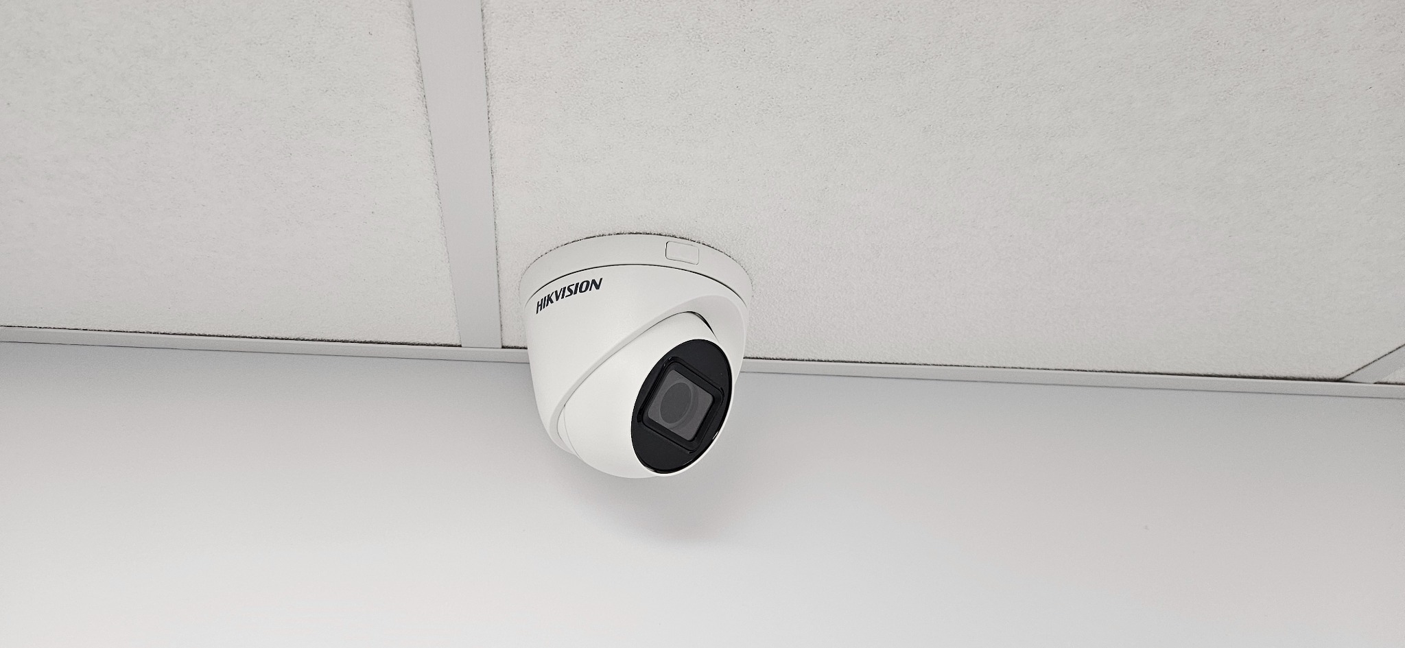 Kamera monitoringu Hikvision, kopułkowa, zamontowana na białym suficie podwieszanym z widocznymi listwami. Obiektyw skierowany w dół.