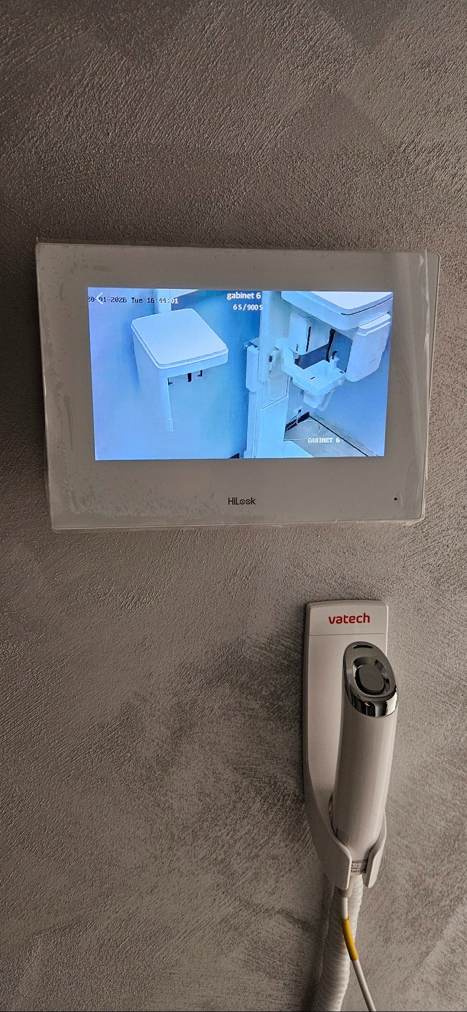 Ekran monitoringu HiLook z widokiem na gabinet stomatologiczny i urządzenie Vatech na ścianie o szarej strukturze. Data i godzina widoczne na ekranie.