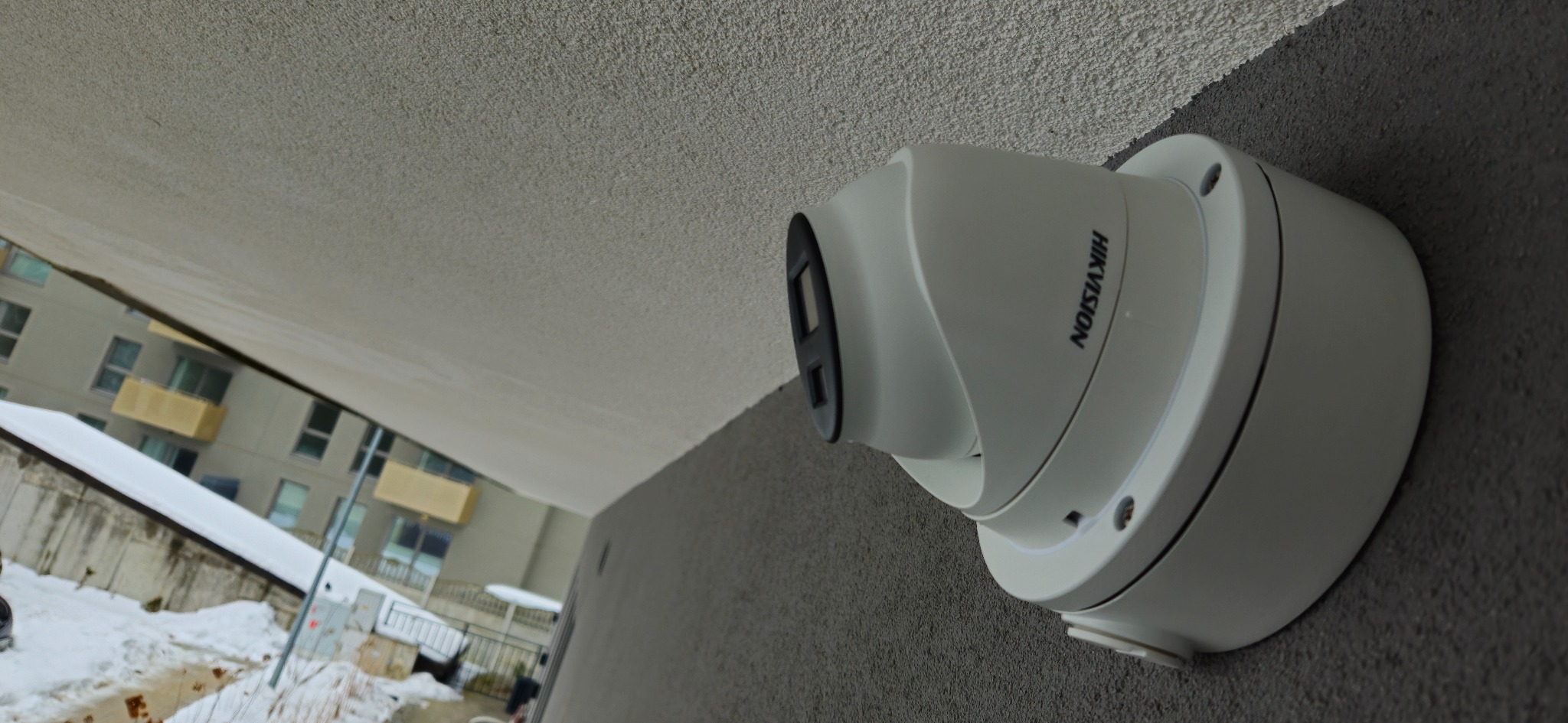 Kamera monitoringu Hikvision, zamontowana na szarej ścianie budynku. Widoczny fragment budynku w tle, z elementami zimowego krajobrazu. Ujęcie z dołu, podkreślające solidność instalacji.