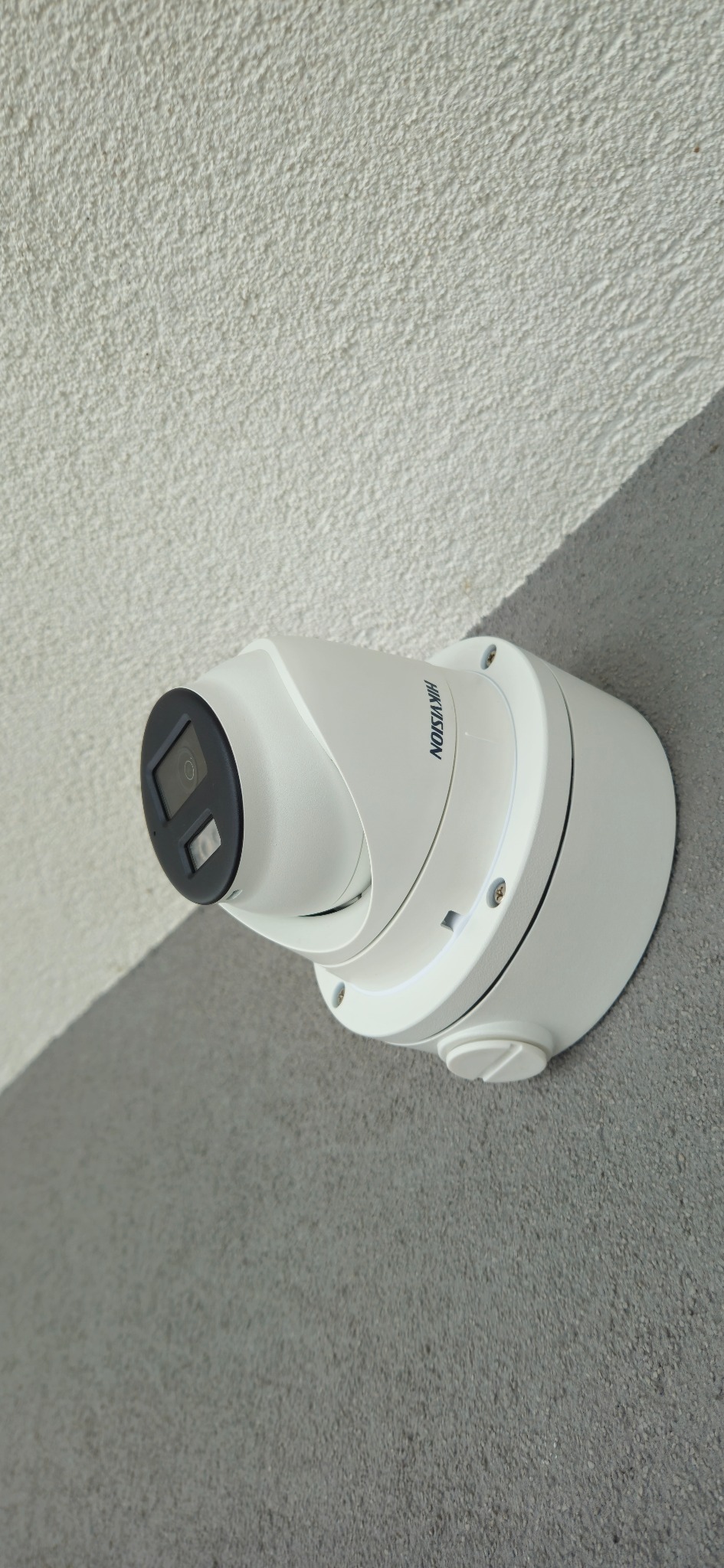 Kamera monitoringu Hikvision, zamontowana na dwukolorowej ścianie z wyraźną fakturą. Widoczne śruby mocujące i obiektyw kamery. Ujęcie z dołu, podkreślające solidność instalacji.
