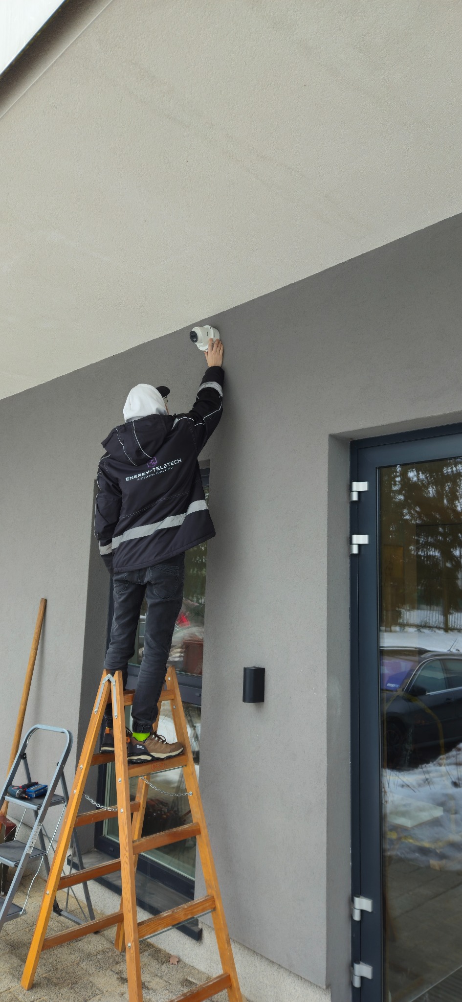Instalator na drabinie montuje kamerę monitoringu na elewacji budynku. Widoczna boczna ściana budynku z oknem i fragmentem zaparkowanego samochodu.